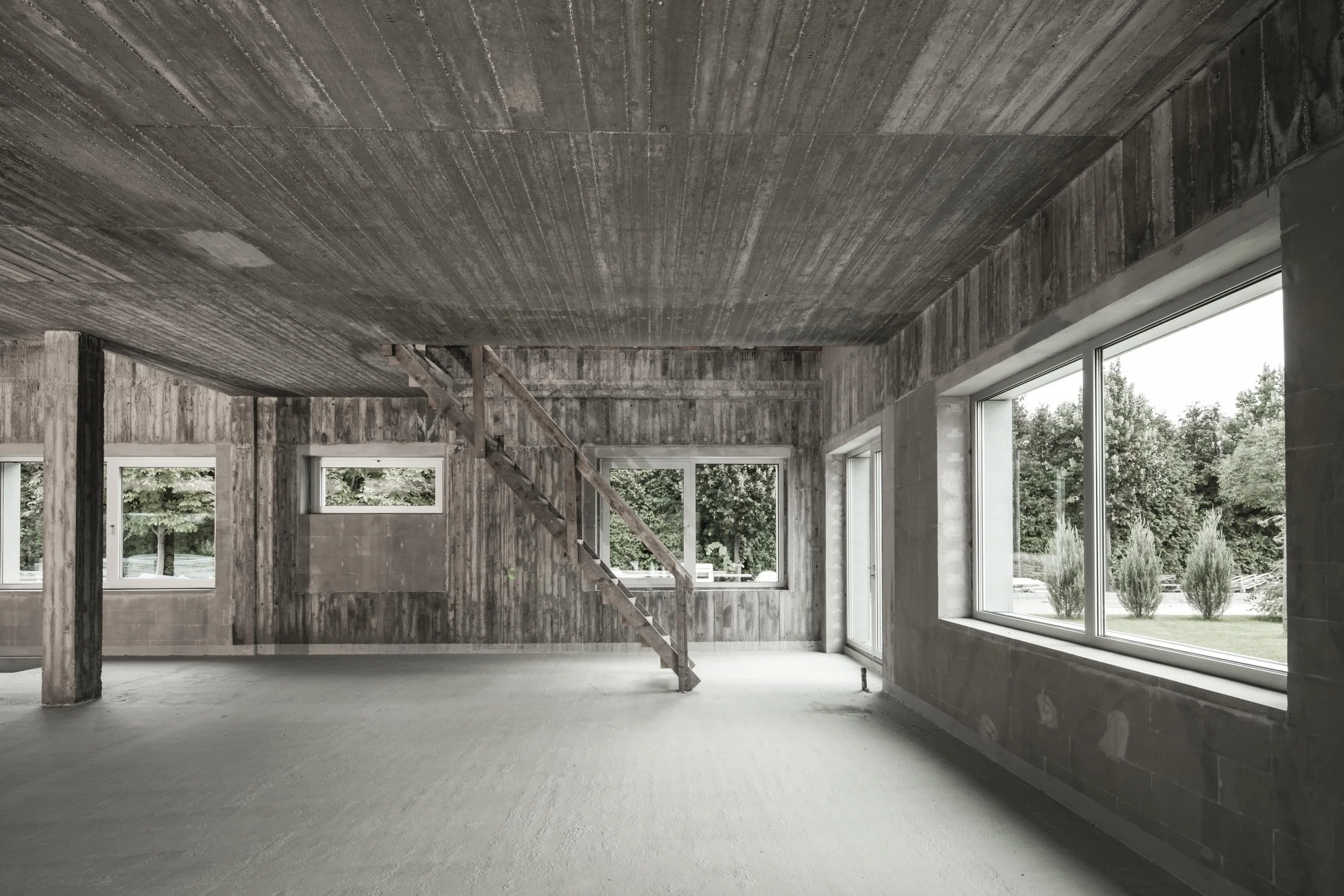 Interior of a home under construction with unfinished walls, ceiling, and floor, featuring large windows showing outdoor greenery, and a wooden staircase leading to an upper level.