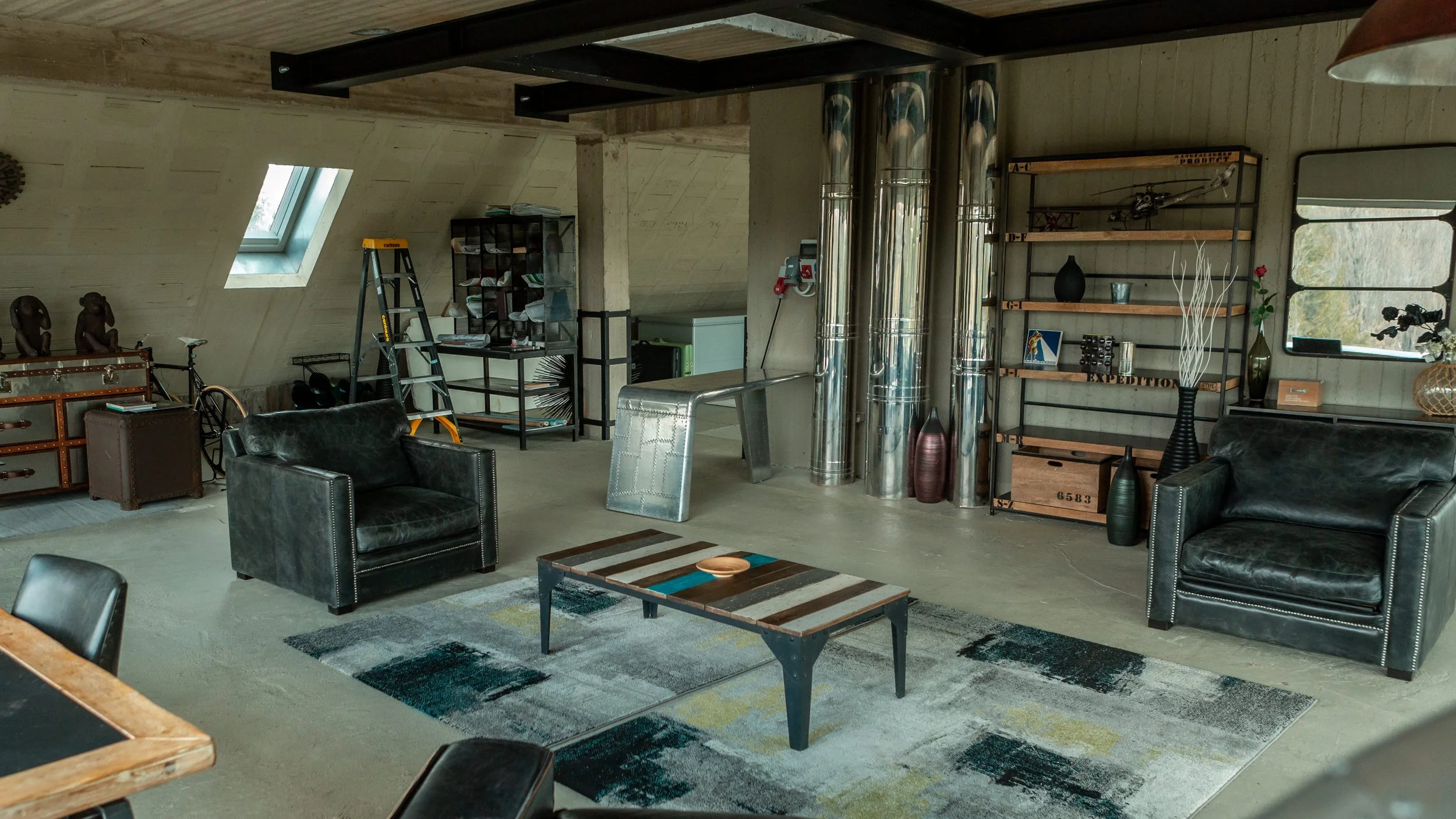 Living room with black leather armchairs, a wooden coffee table, metal shelving with various decorative items, a ladder, and metallic pipes in a spacious, modern style.