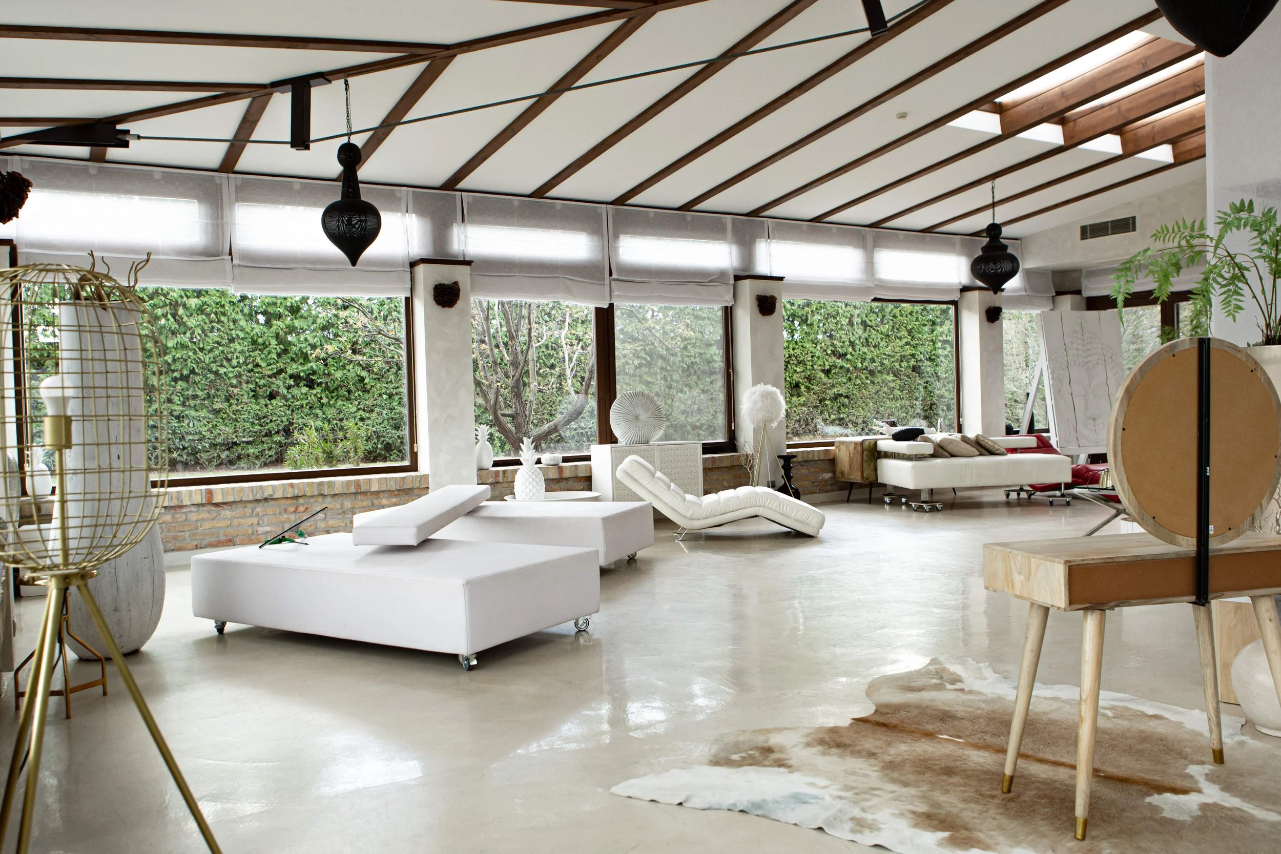 Spacious interior room with large windows showing trees outside, modern white furniture including sofas, lounge chair, and a cowhide rug, with decorative plants and art pieces.
