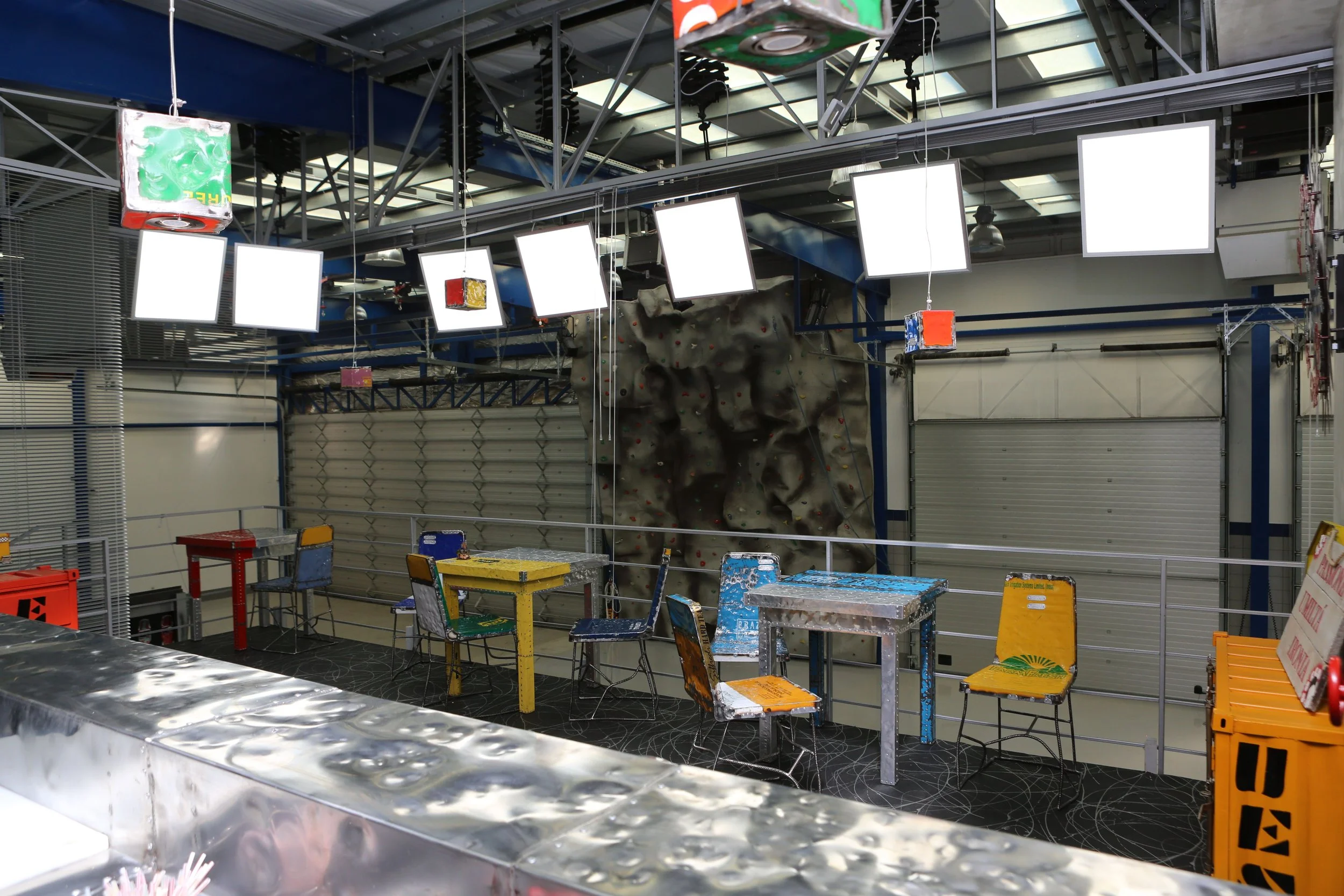 Indoor rock climbing gym with a climbing wall in the background and colorful tables and chairs in the foreground.