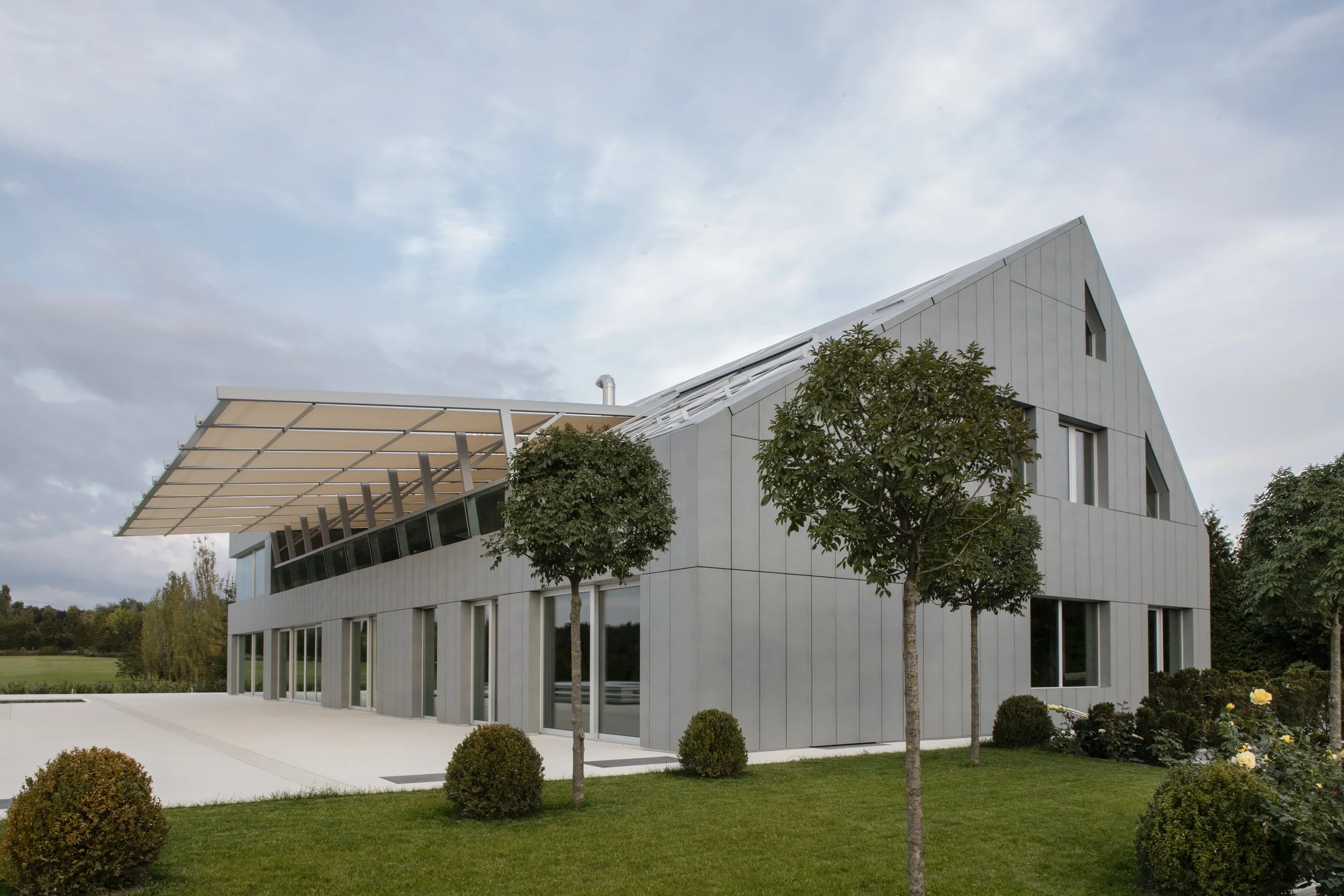 Modern building with sloped roof, large windows, and surrounding landscaped lawn with trees and bushes.