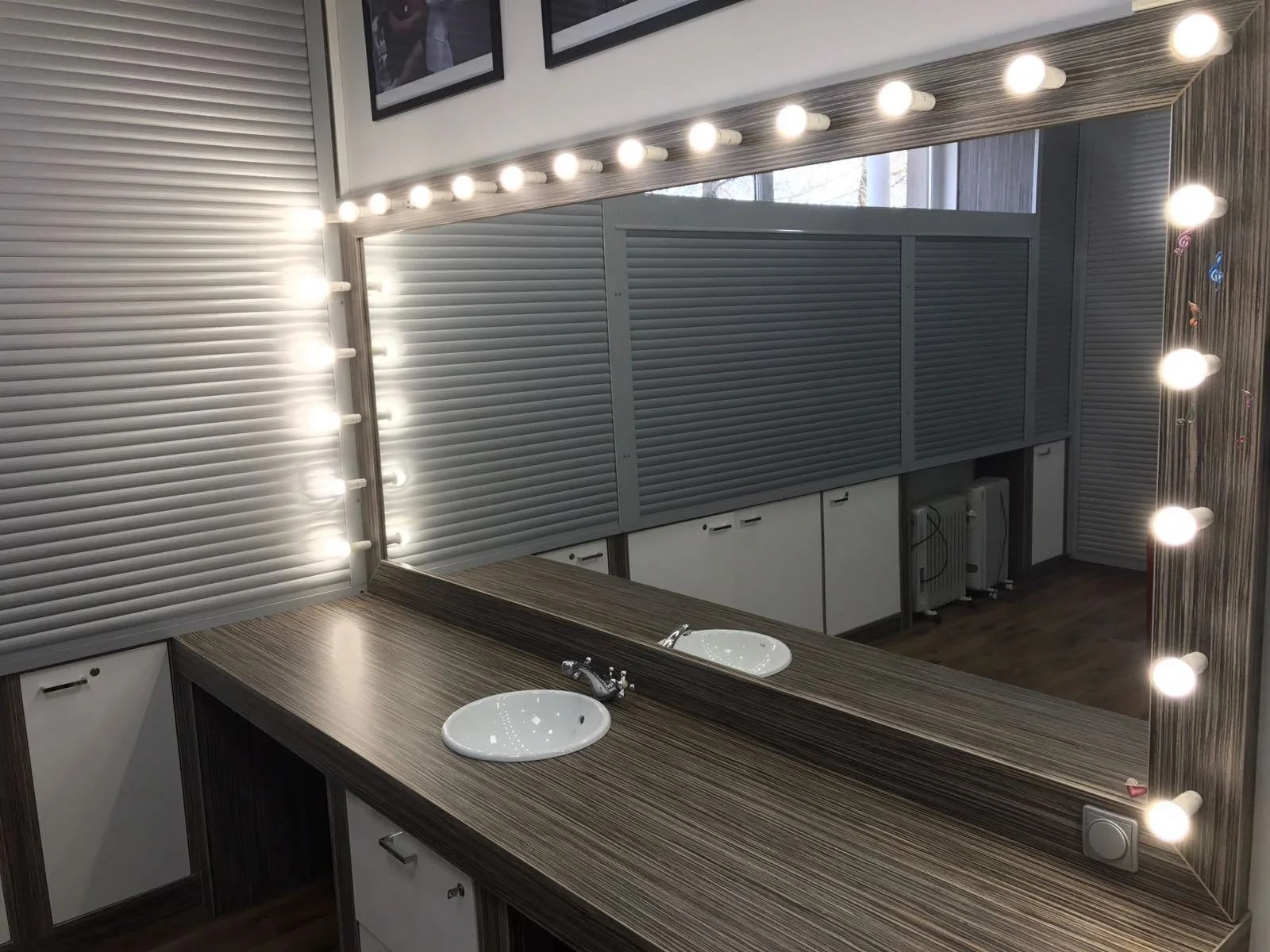 A large rectangular mirror with exposed light bulbs around the edges, mounted on a wall with two small sinks below it in a bathroom or dressing room.