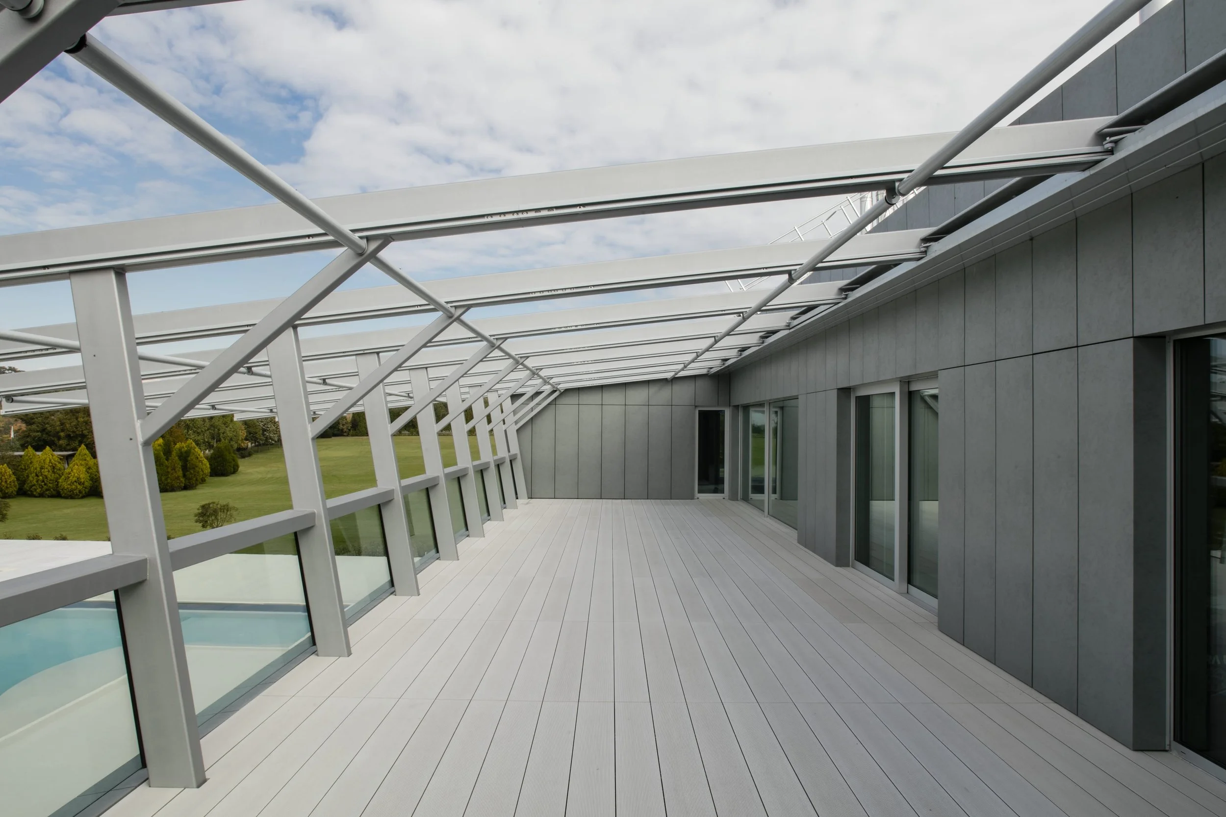 Empty modern balcony with glass railings and a retractable awning, attached to a gray building with large windows, overlooking a green lawn and trees under a partly cloudy sky.