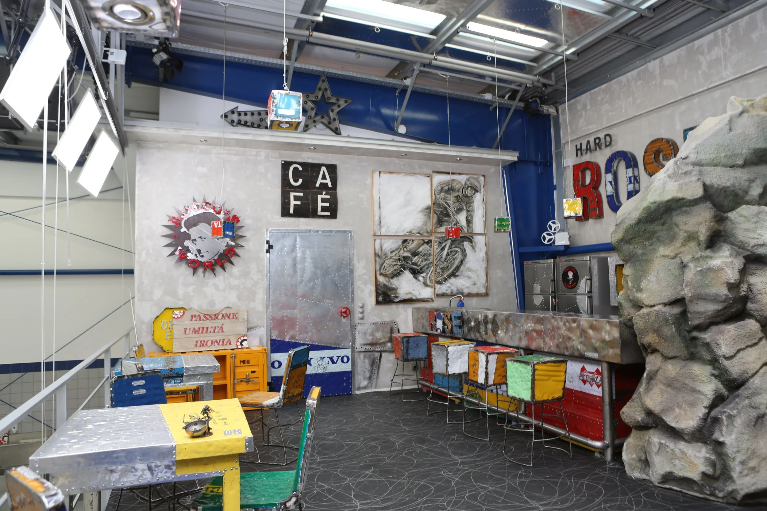 Inside a cafe decorated with colorful metal art, a rock climbing wall, and vintage furniture, with words like 'CAFE' and 'ROS' visible on the walls and vibrant hanging lights.