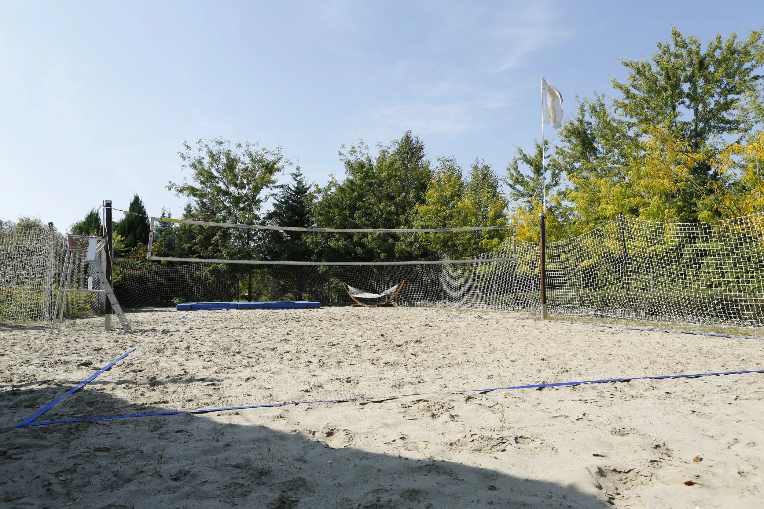 A sand volleyball court with a net, a volleyball, and a lifeguard chair on the left, surrounded by trees with green leaves under a clear blue sky.