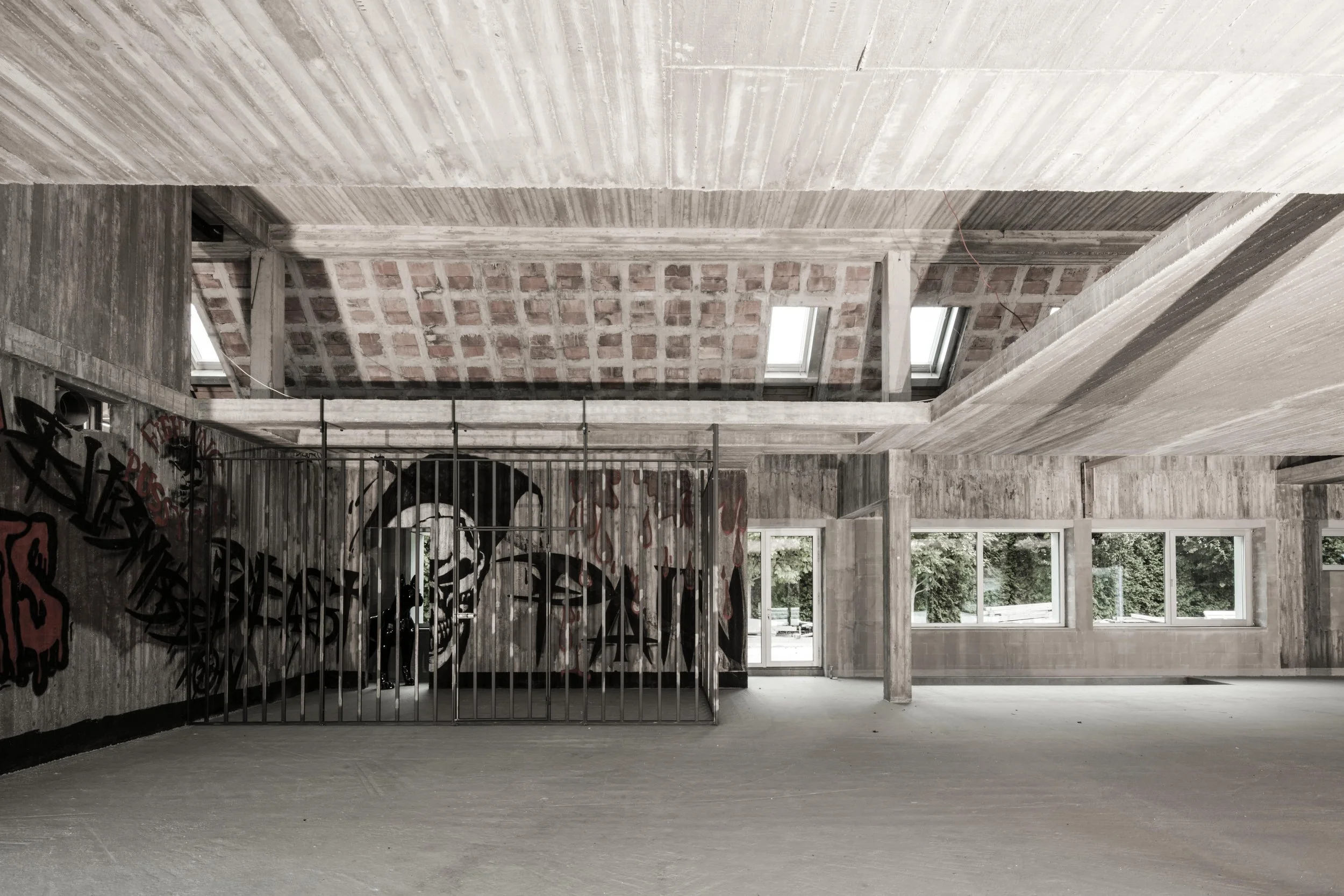Unfinished, urban interior space with concrete walls and ceiling, large windows, graffiti on a partition, and skylights in the ceiling.