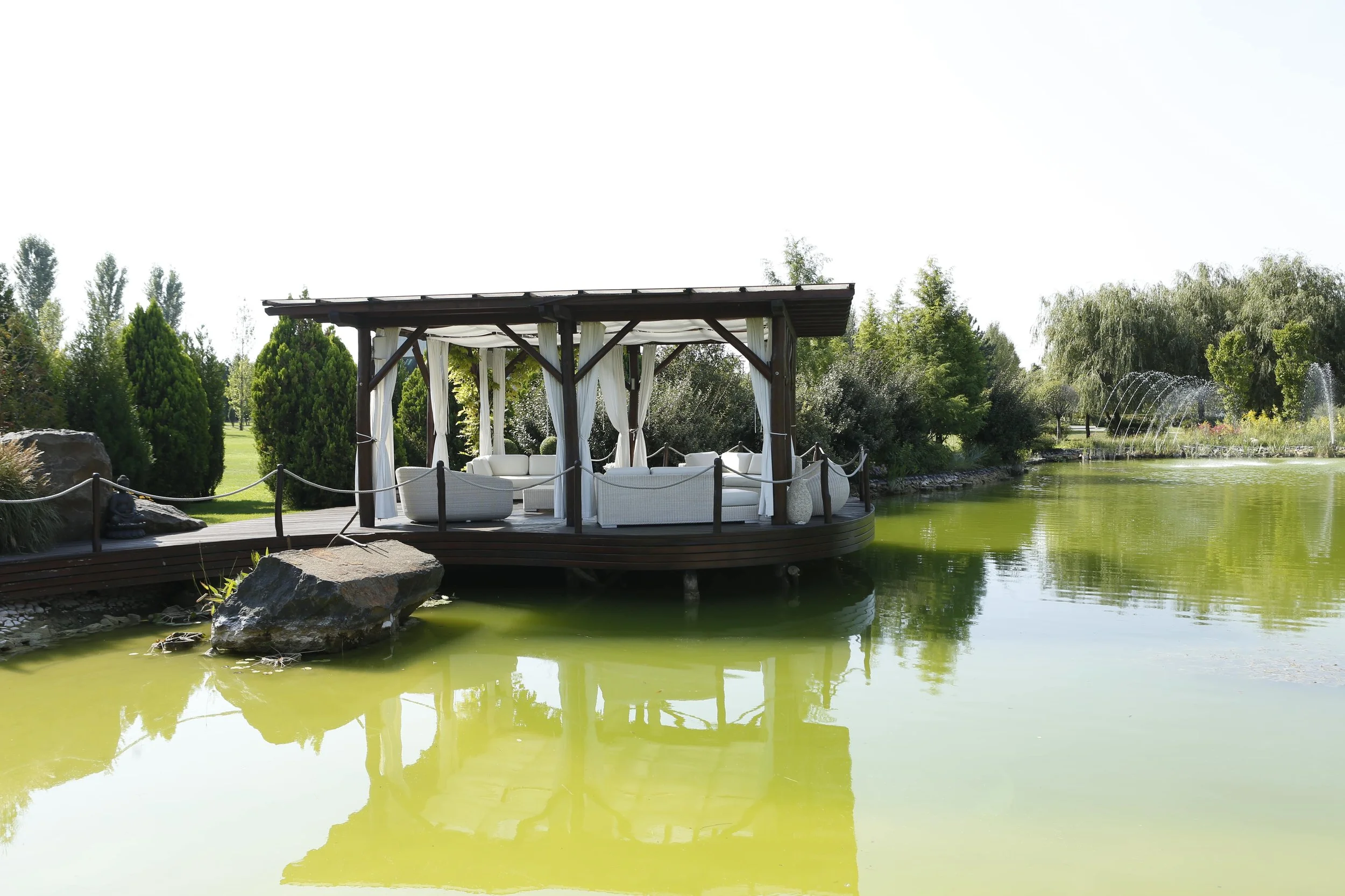 A wooden gazebo with white curtains and outdoor furniture, next to a pond with greenish water, trees, shrubs, and a fountain in the background.