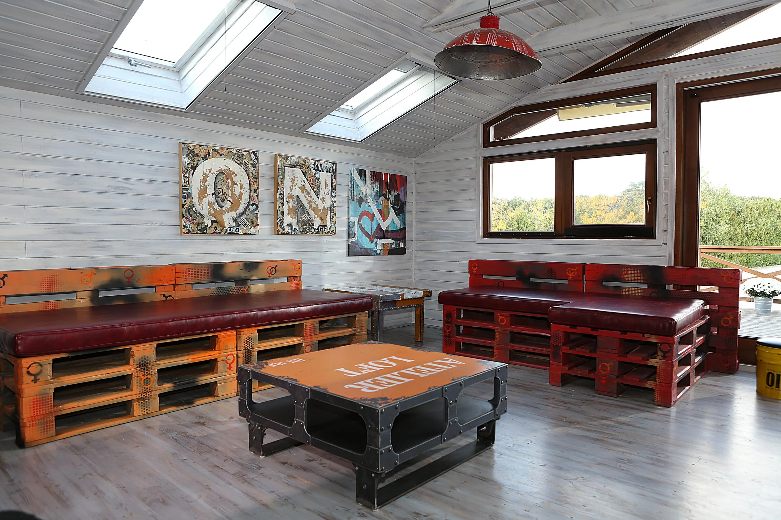 Living room with two pallet sofas, a pallet coffee table, and wall art, featuring large windows and skylights in a rustic style.