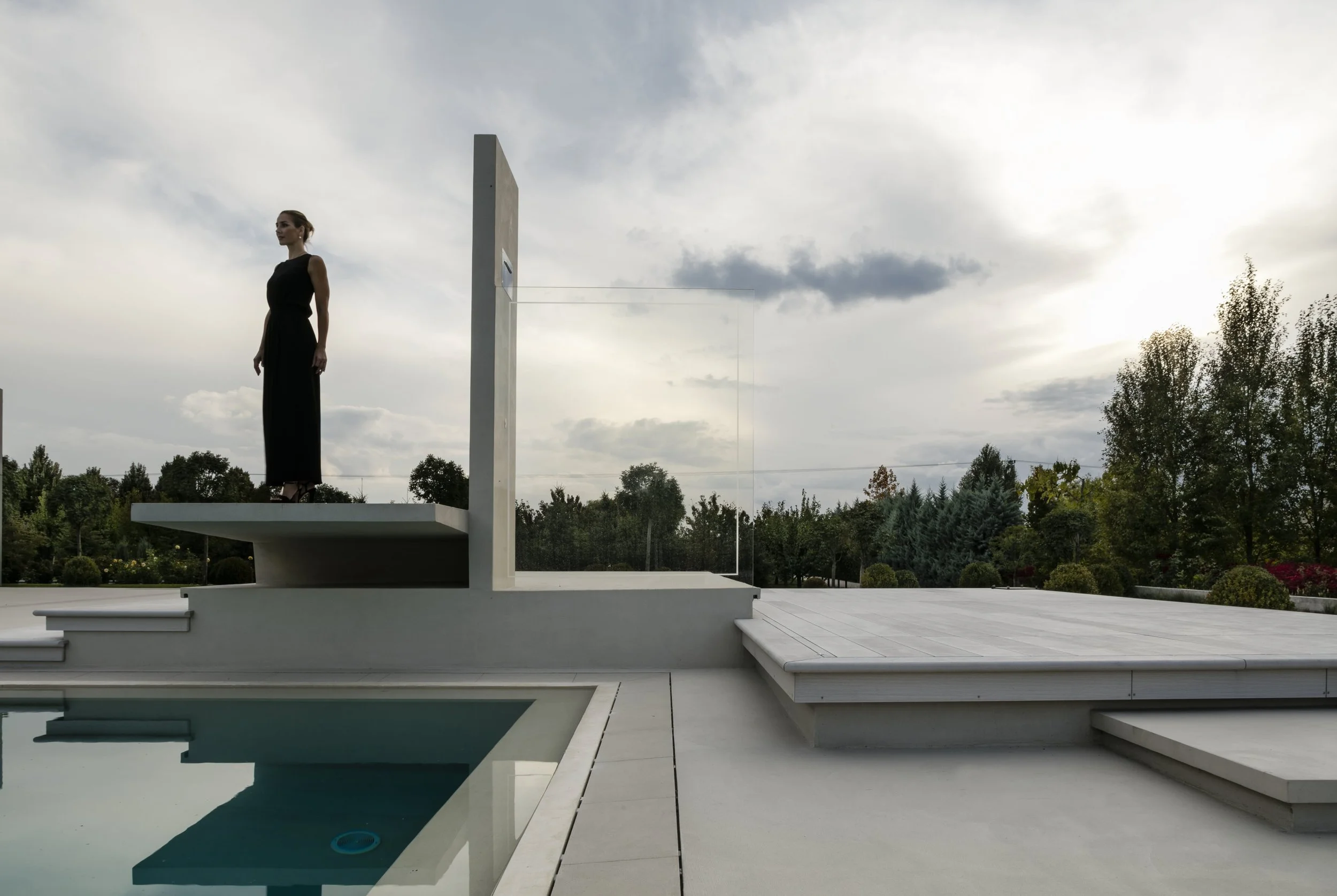 Woman in a black dress standing on a modern white platform near a pool, outdoors with trees and cloudy sky in background.