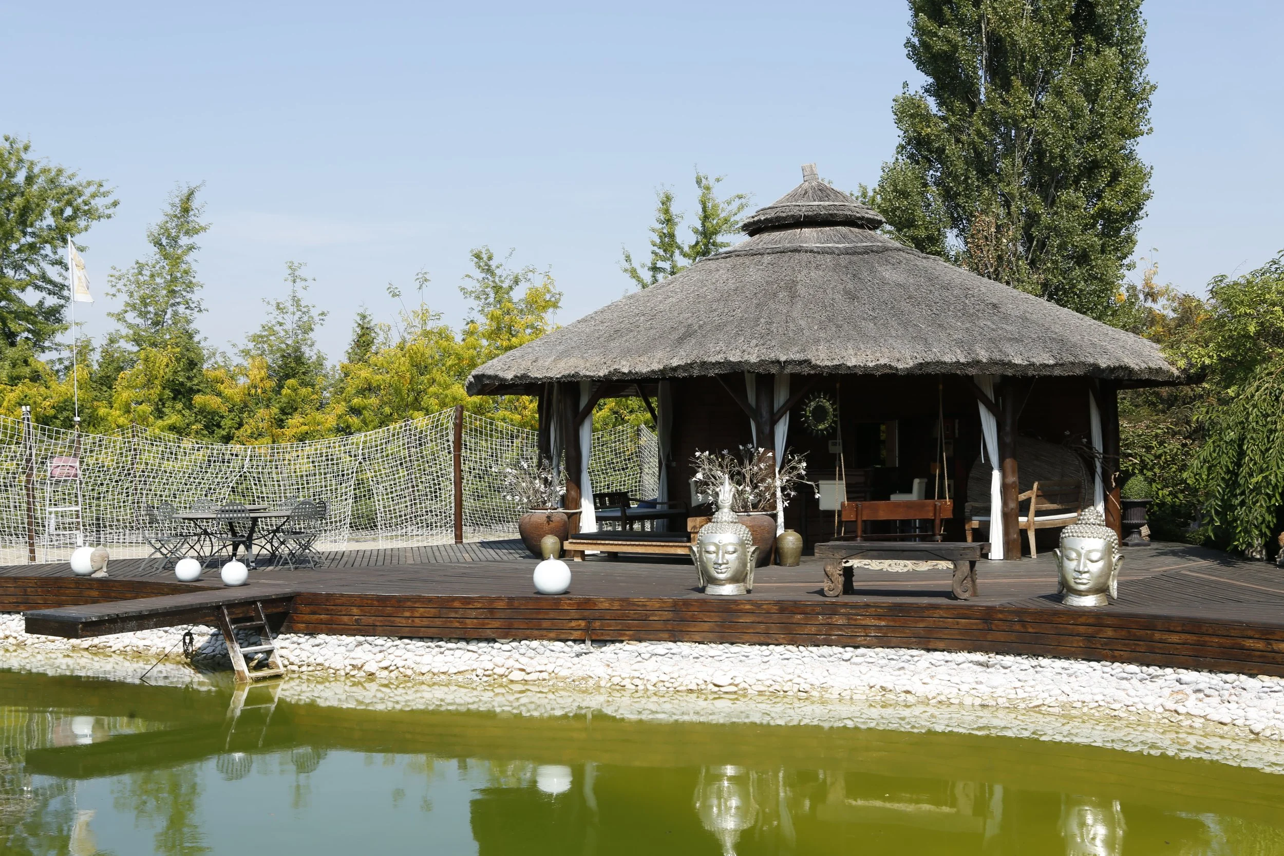 Outdoor scene with a thatched-roof gazebo on a wooden deck by a pond, decorated with large Buddha head sculptures and vases, surrounded by greenery and trees.