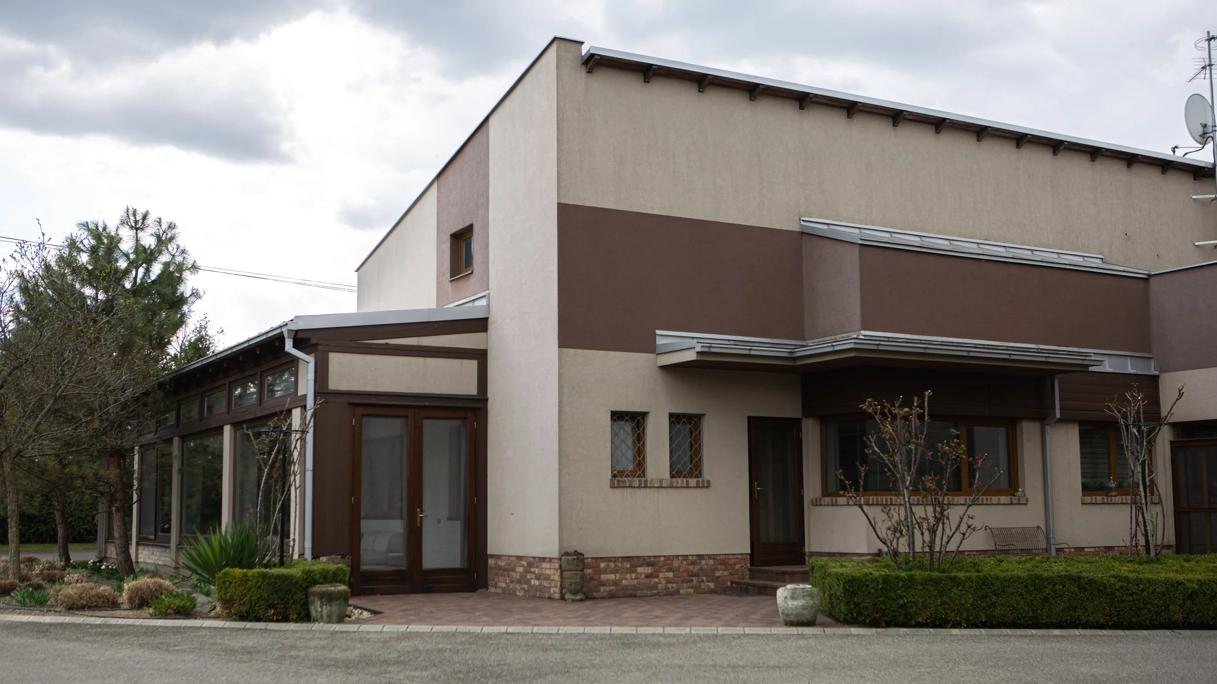 A two-story residential house with a beige and brown exterior, featuring several windows, an enclosed porch area with glass and wood frames, small landscaped bushes and trees in front, and a paved walkway leading to the doors.