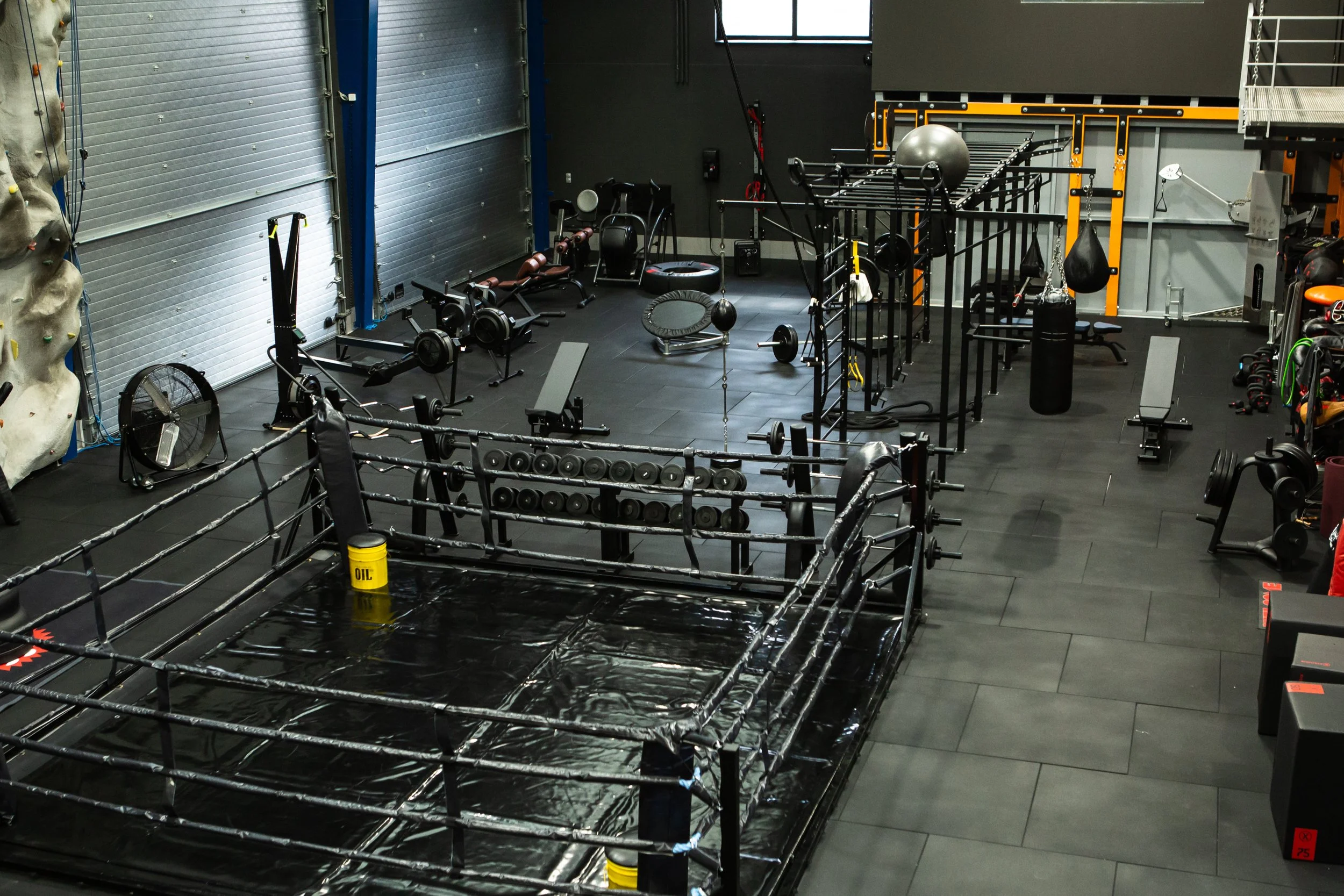 Empty boxing gym featuring a boxing ring in the foreground, various exercise equipment on black flooring, including weights, benches, a punching bag, and a climbing wall on the left side.