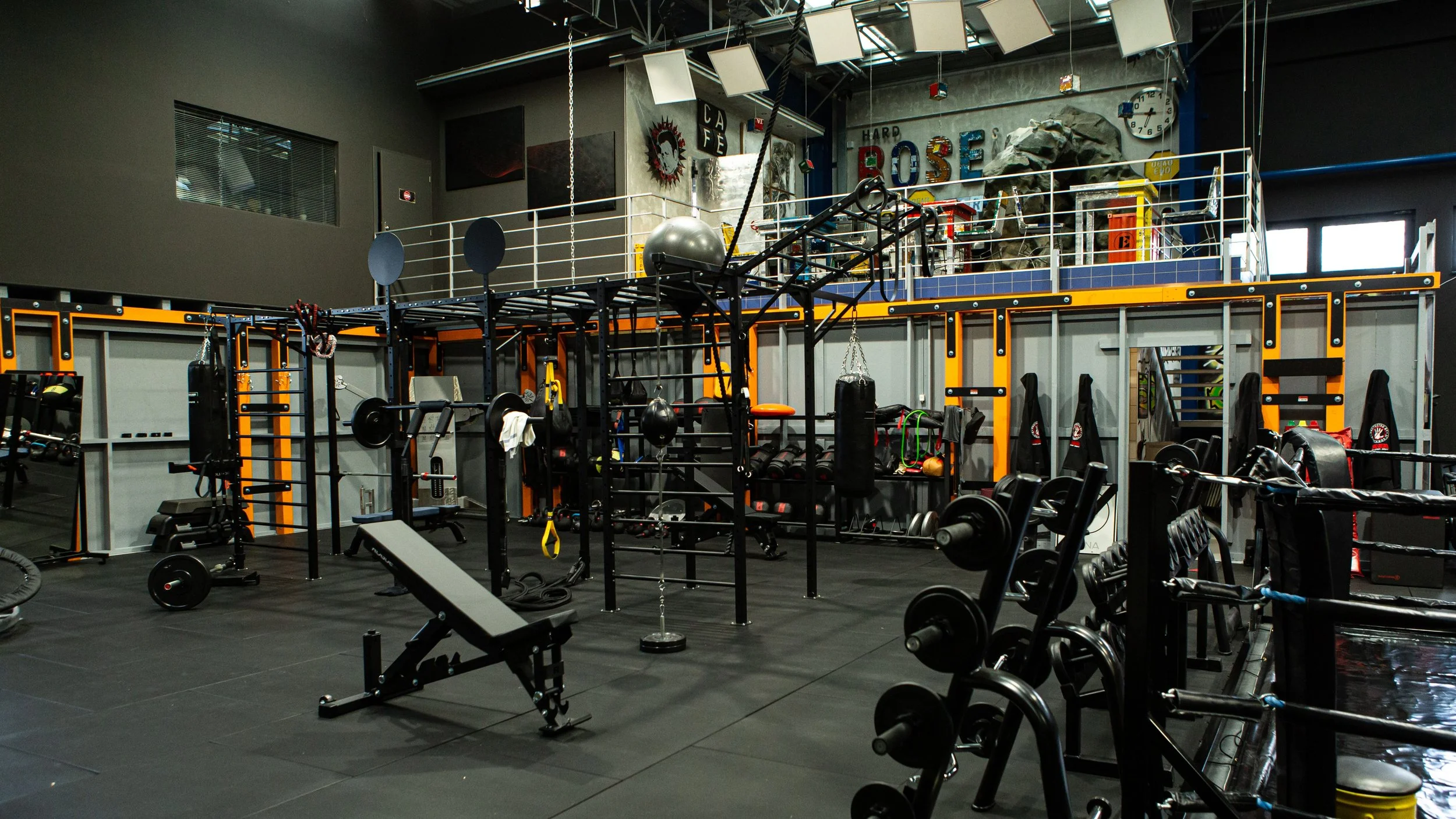 Inside a modern gym with various exercise equipment, weights, and a large climbing and training structure. The background features a wall with colorful decorations and a large clock.