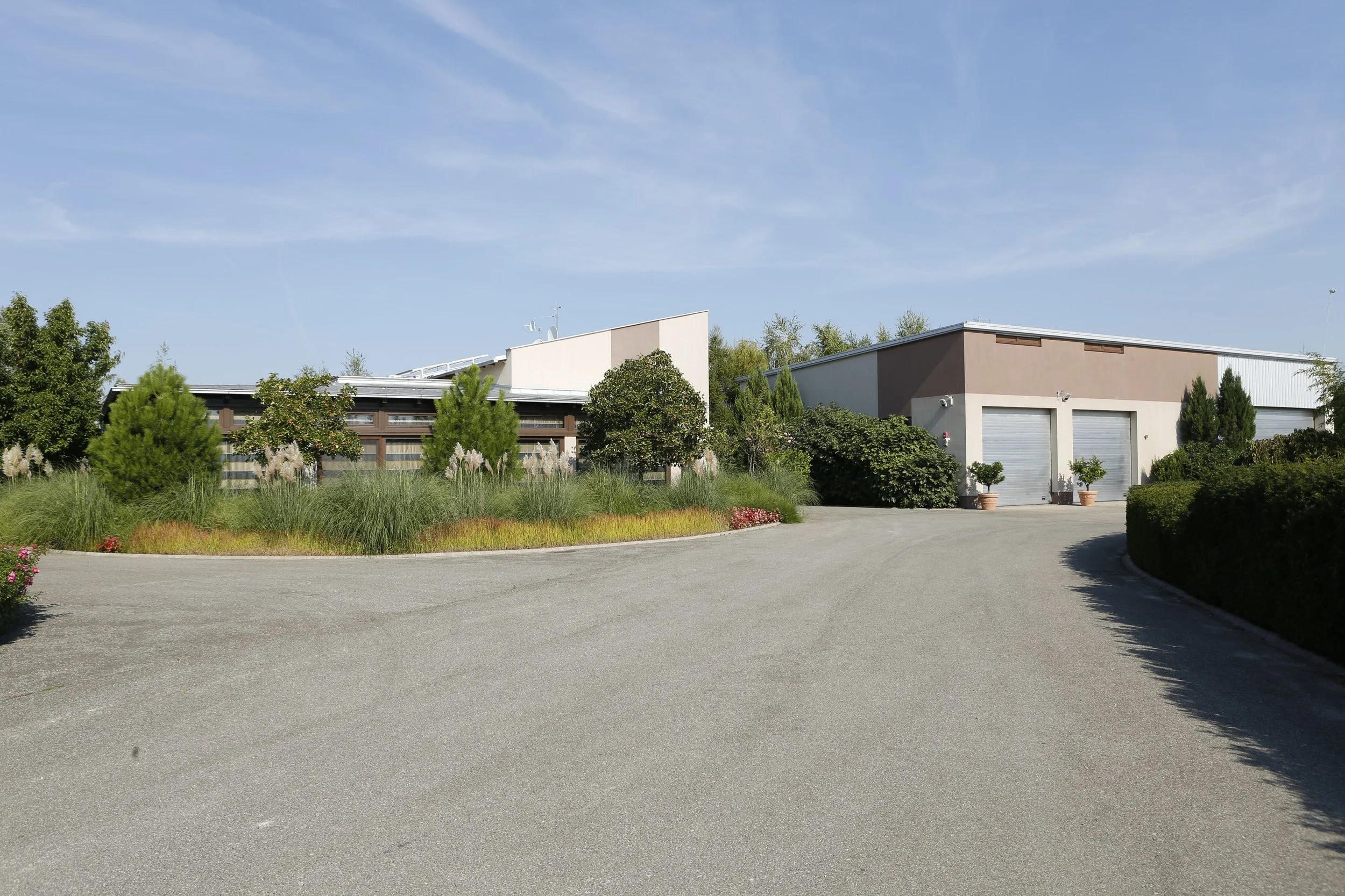 A winding driveway leading up to a modern warehouse or garage with metal roll-up doors, surrounded by landscaped greenery and trees under a clear blue sky.