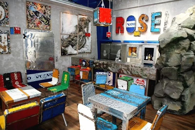Interior of a restaurant or bar decorated with artwork and colorful furniture made from recycled materials, including a bar counter, chairs, and large decorative letters spelling 'ROSE' on the wall.