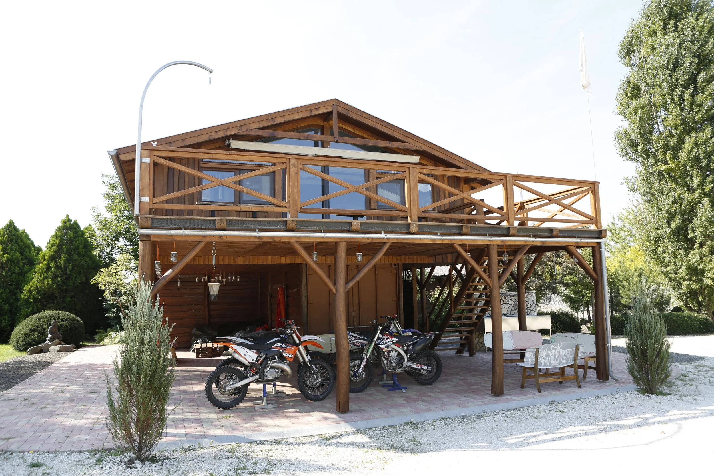 Two motorcycles parked under a wooden house with a deck, trees in the background, and outdoor furniture.
