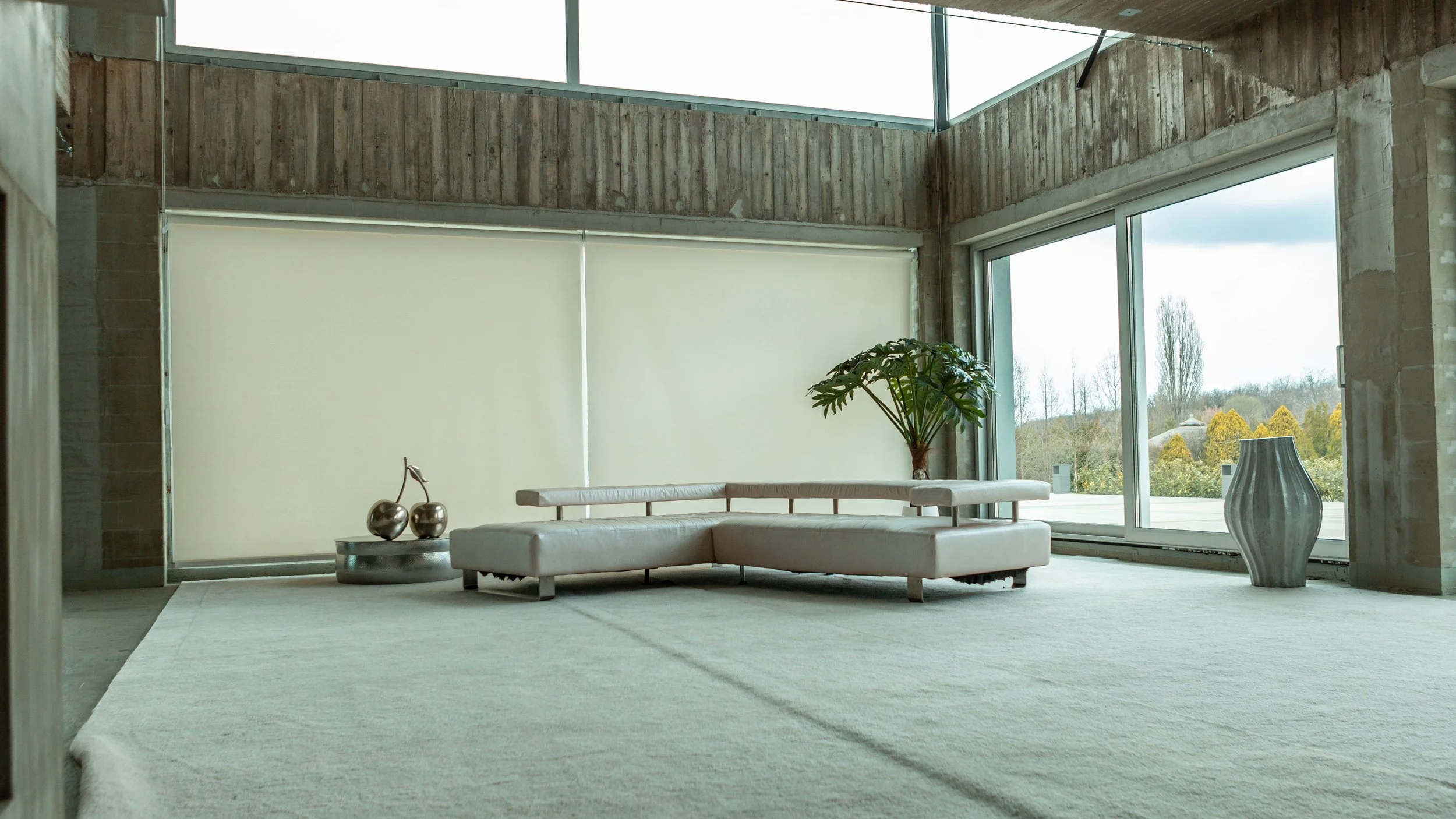 Modern living room with a beige sectional sofa, large windows with views of trees, potted plant, decorative vases, and minimalist decor.