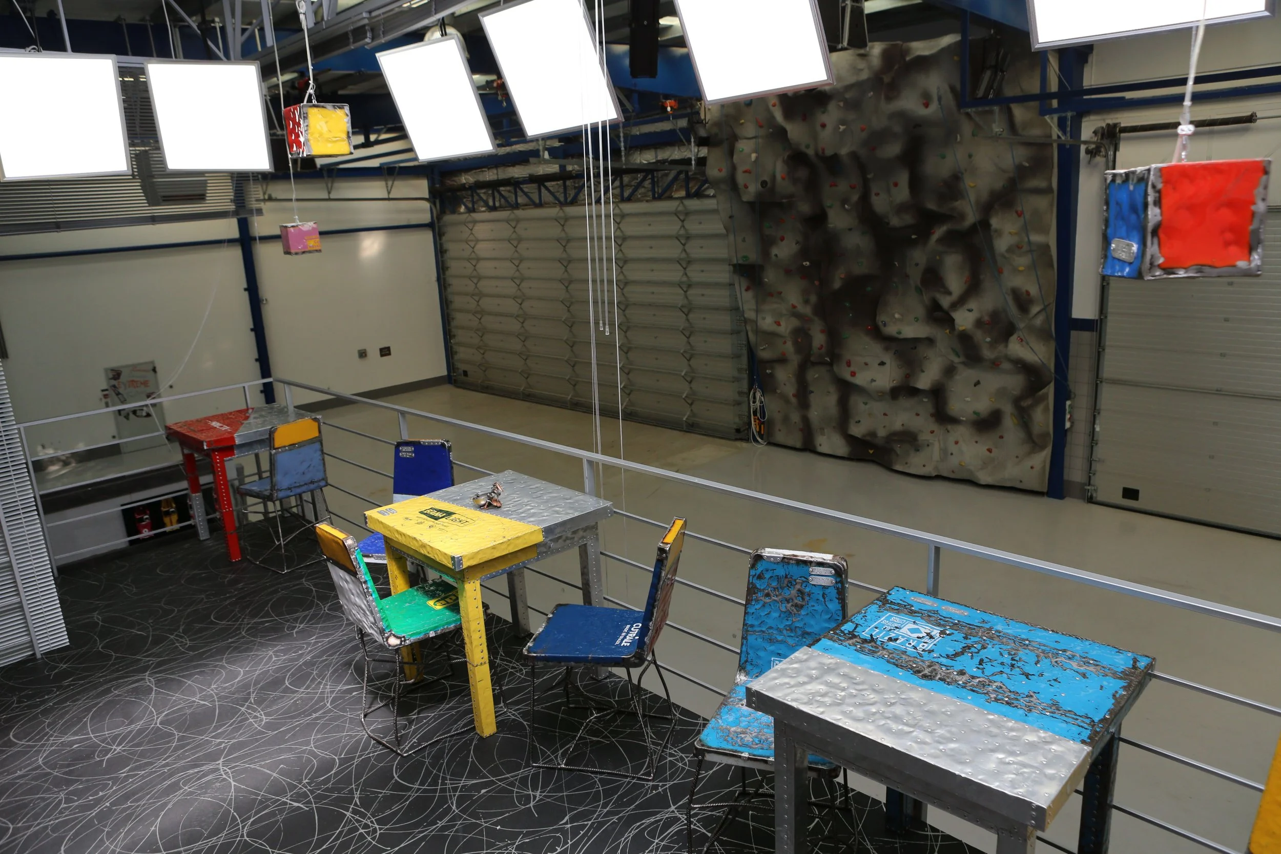 Indoor rock climbing gym with a large climbing wall on the wall and a foreground seating area with colorful metal tables and chairs, and multiple flat-screen monitors hanging from the ceiling.