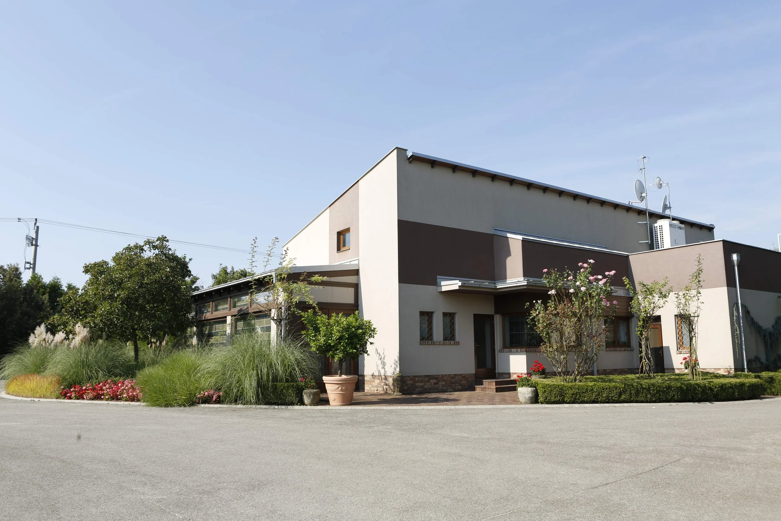 Modern two-story house with beige and brown exterior, surrounded by landscaped greenery including trees, bushes, and flowering plants, under a clear blue sky.