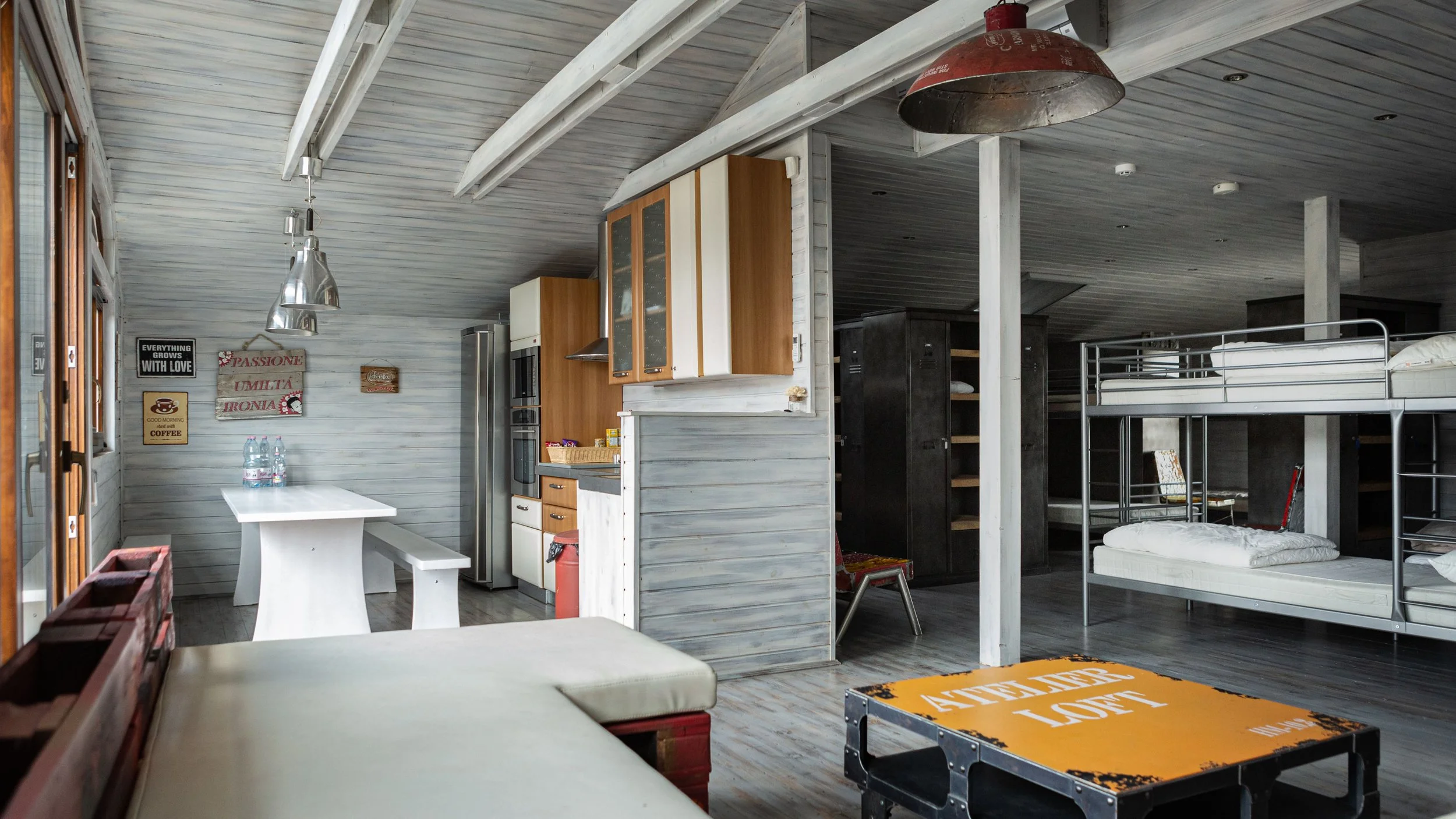 A spacious, rustic-style room with wooden walls and ceiling, containing bunk beds, a small kitchenette, a dining area with bottles of water on the table, and various wall signs and decorations.