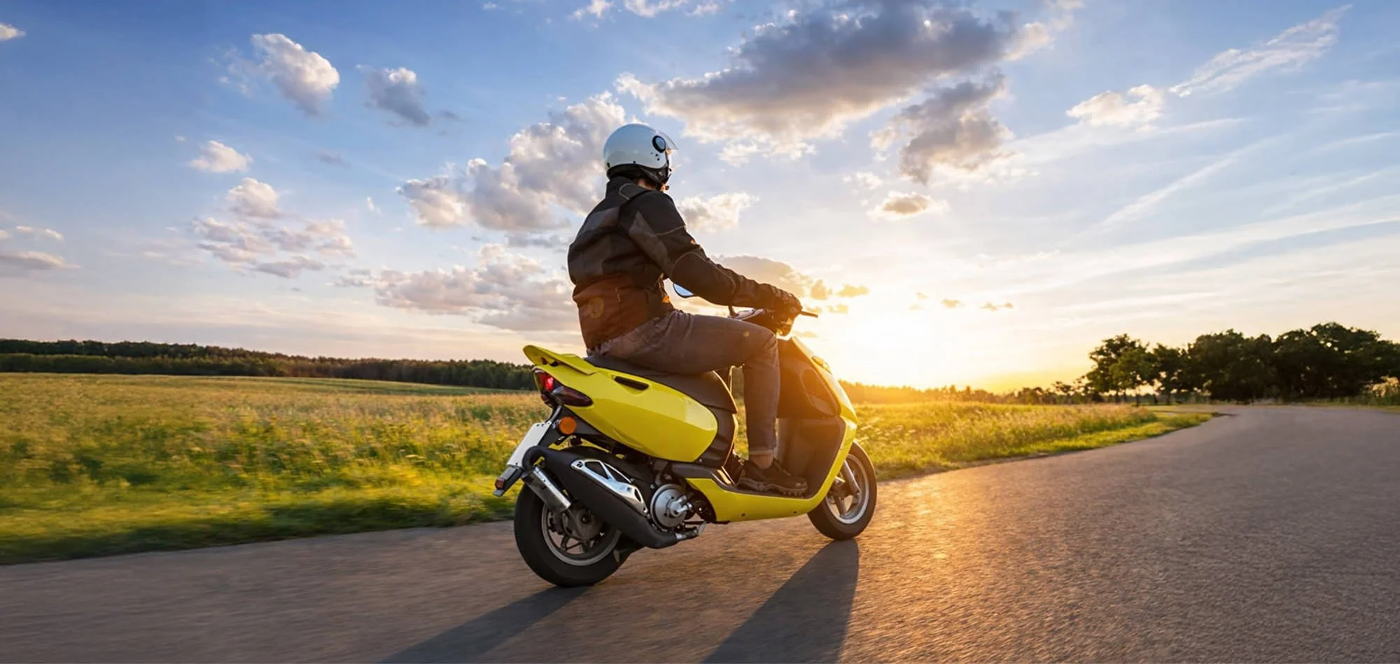 Person riding a yellow scooter on a curved rural road during sunset
