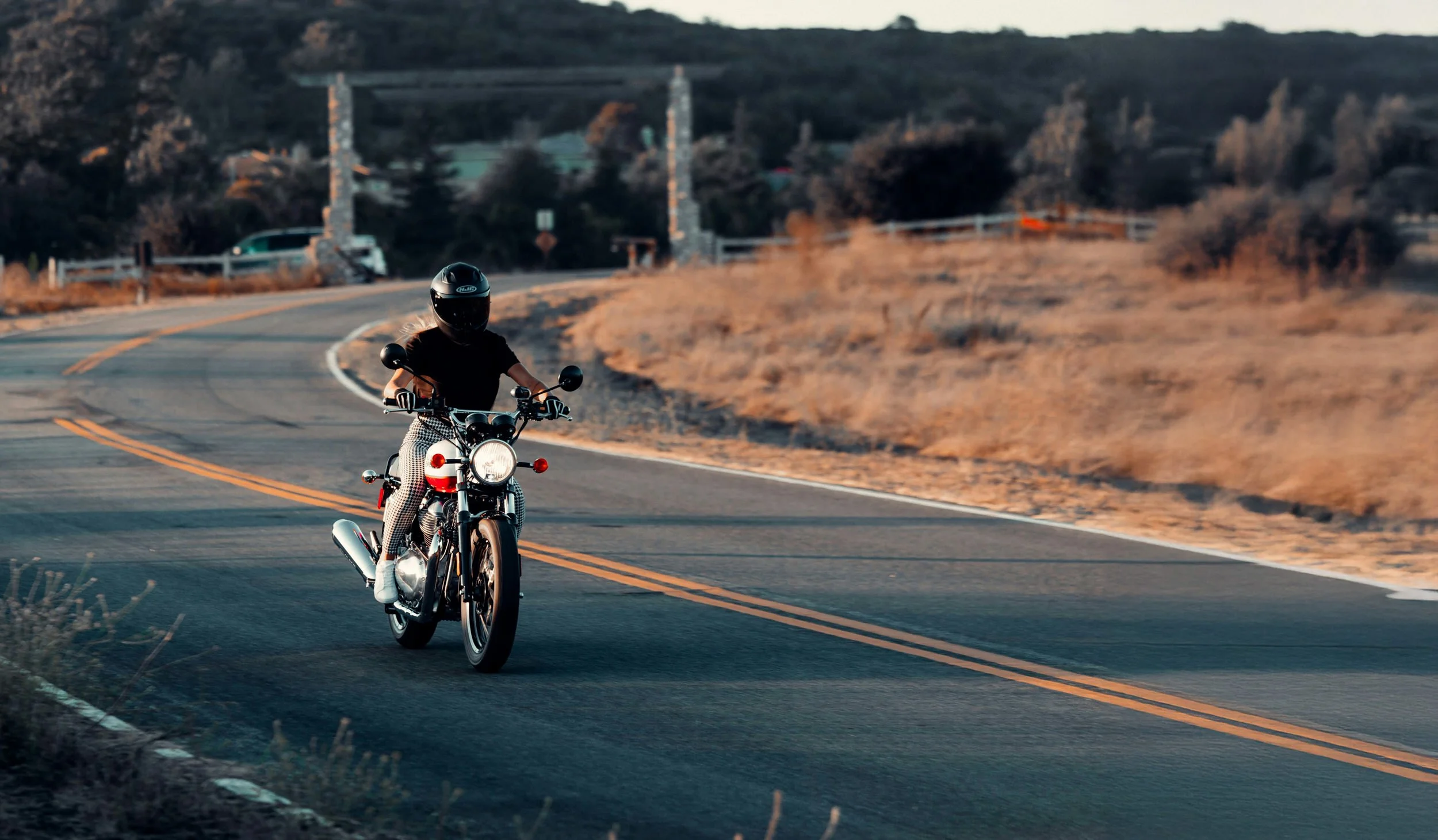 A person riding a motorcycle on a winding rural road during sunset, dressed in black with a black helmet, surrounded by dry grass and sparse trees.