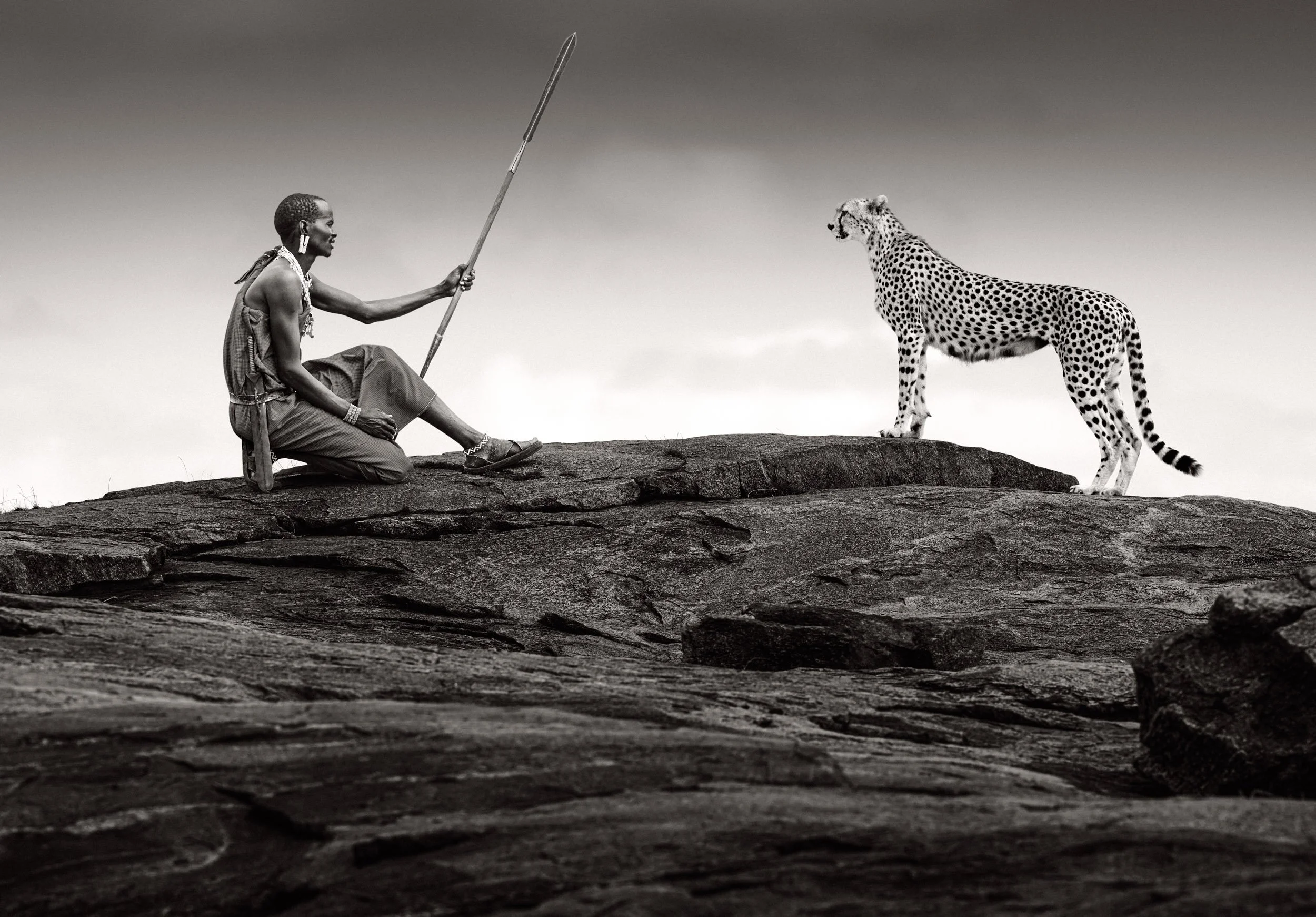 In this composite, a Maasai warrior stands opposite a cheetah on top of a rocky kopje, the two poised against dramatic skies across the African savanna.