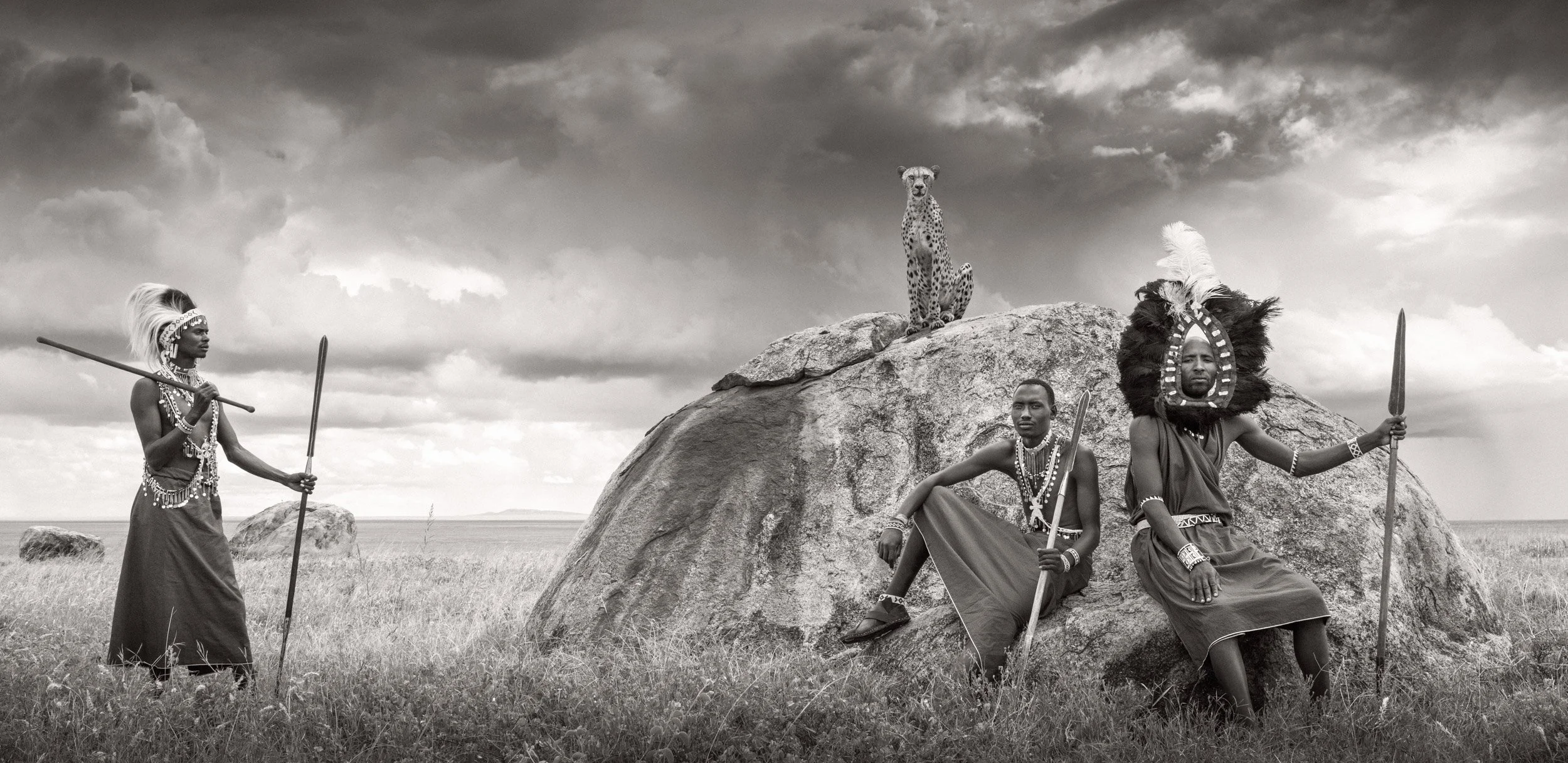 In this composite, three Maasai warriors stand and sit on rocks while a cheetah perches on a kopje behind them beneath dark storm clouds over the African open plains.