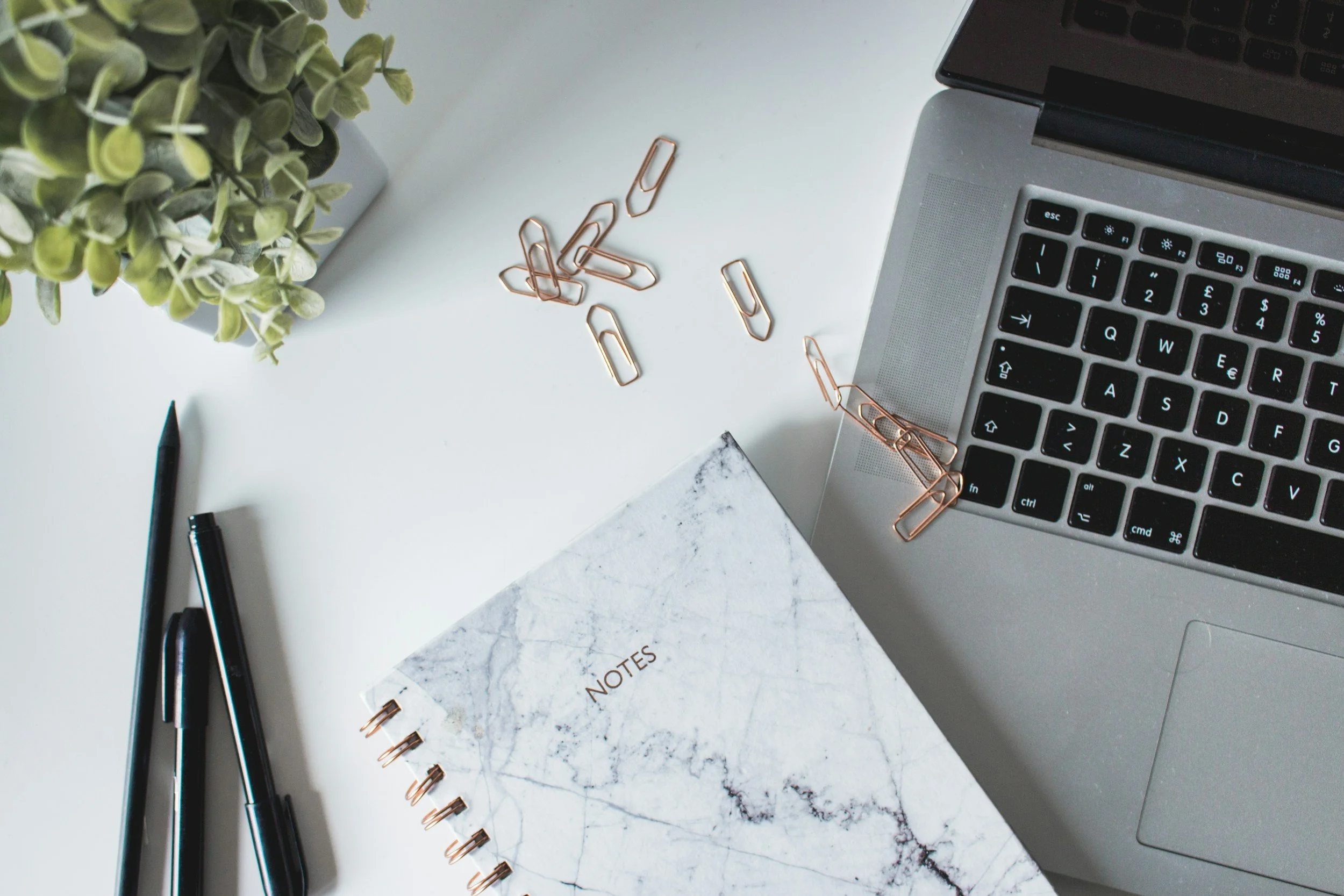 Top view of a workspace with a MacBook, a marble notebook labeled 'NOTES', two black pens, a green plant, and paper clips on a white surface.