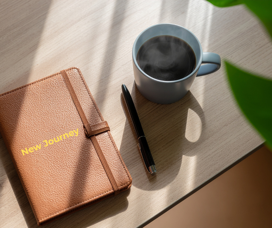 A beige textured journal with yellow text that says "New Journey," a black pen, and a blue coffee mug filled with black coffee on a light-colored wooden table, with shadows and a green plant leaf in the foreground.