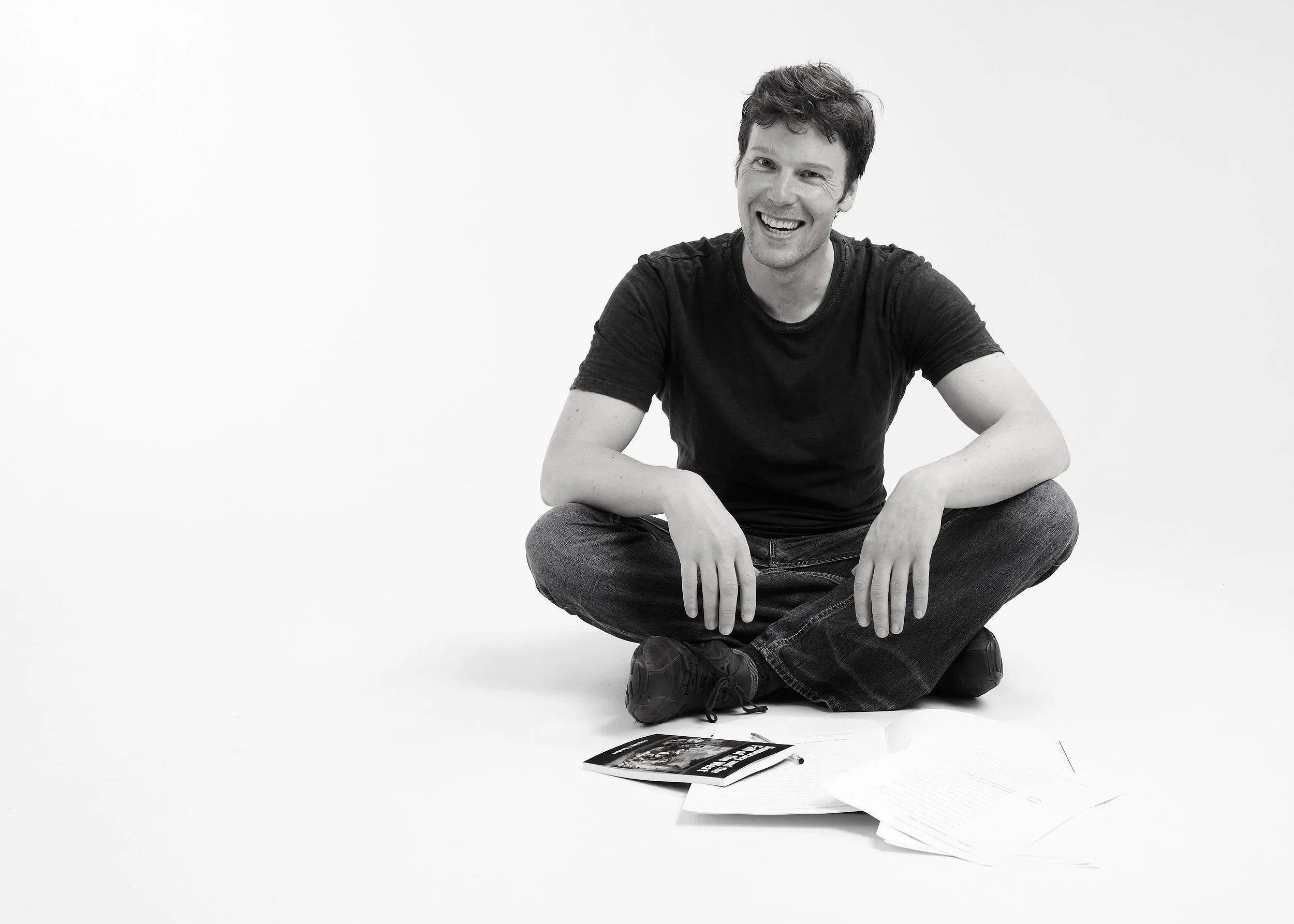 A man sitting cross-legged on the floor, smiling, with papers and a book in front of him.