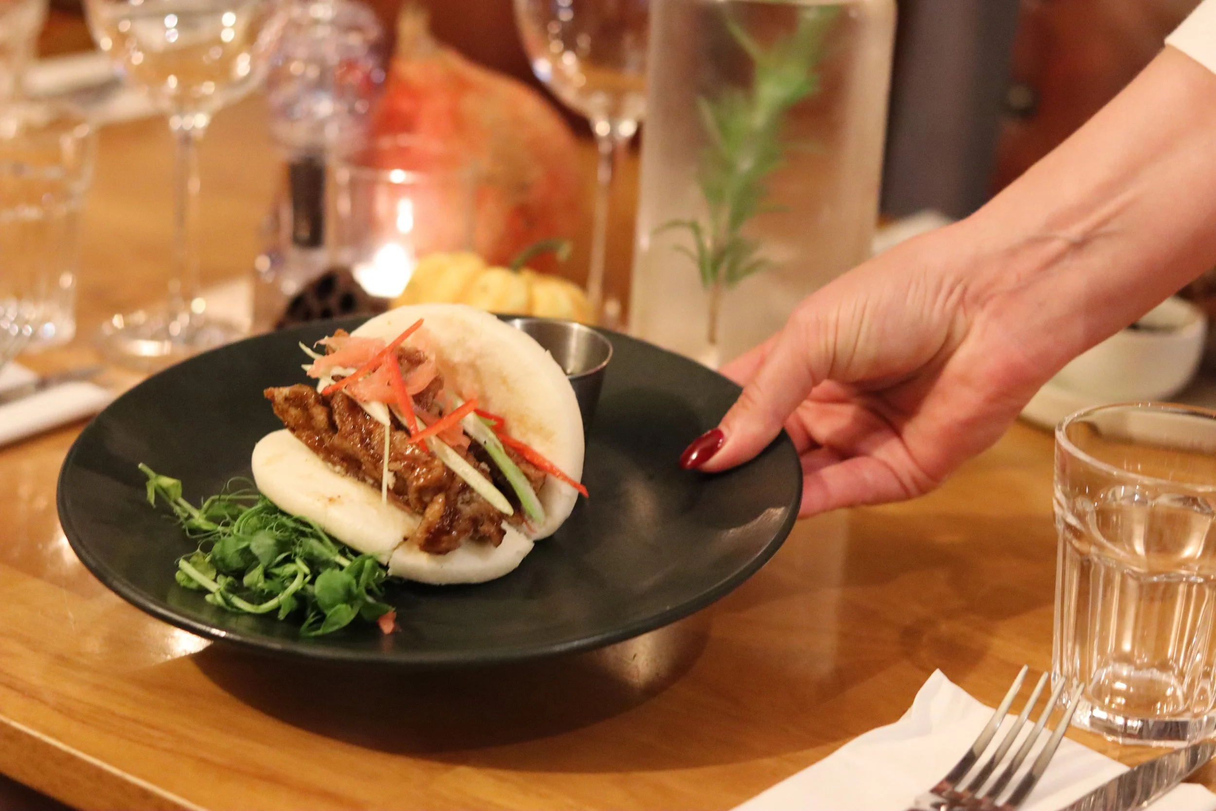 A steamed bun filled with meat, garnished with sliced vegetables, served on a black plate with microgreens, on a wooden table with glasses of water and decorative items in the background.