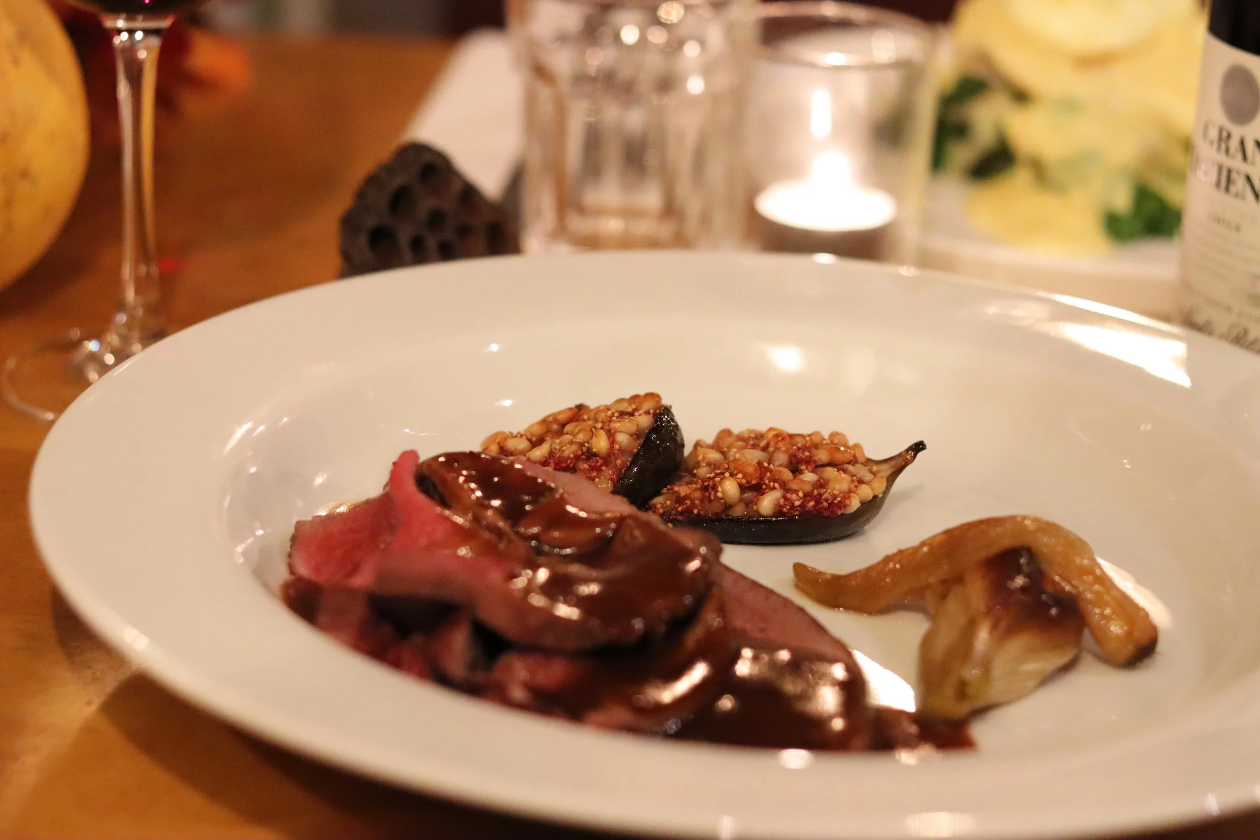 A white plate with slices of roasted meat topped with brown gravy, two grilled fig halves with seeds, and a small roasted banana on a wooden table.