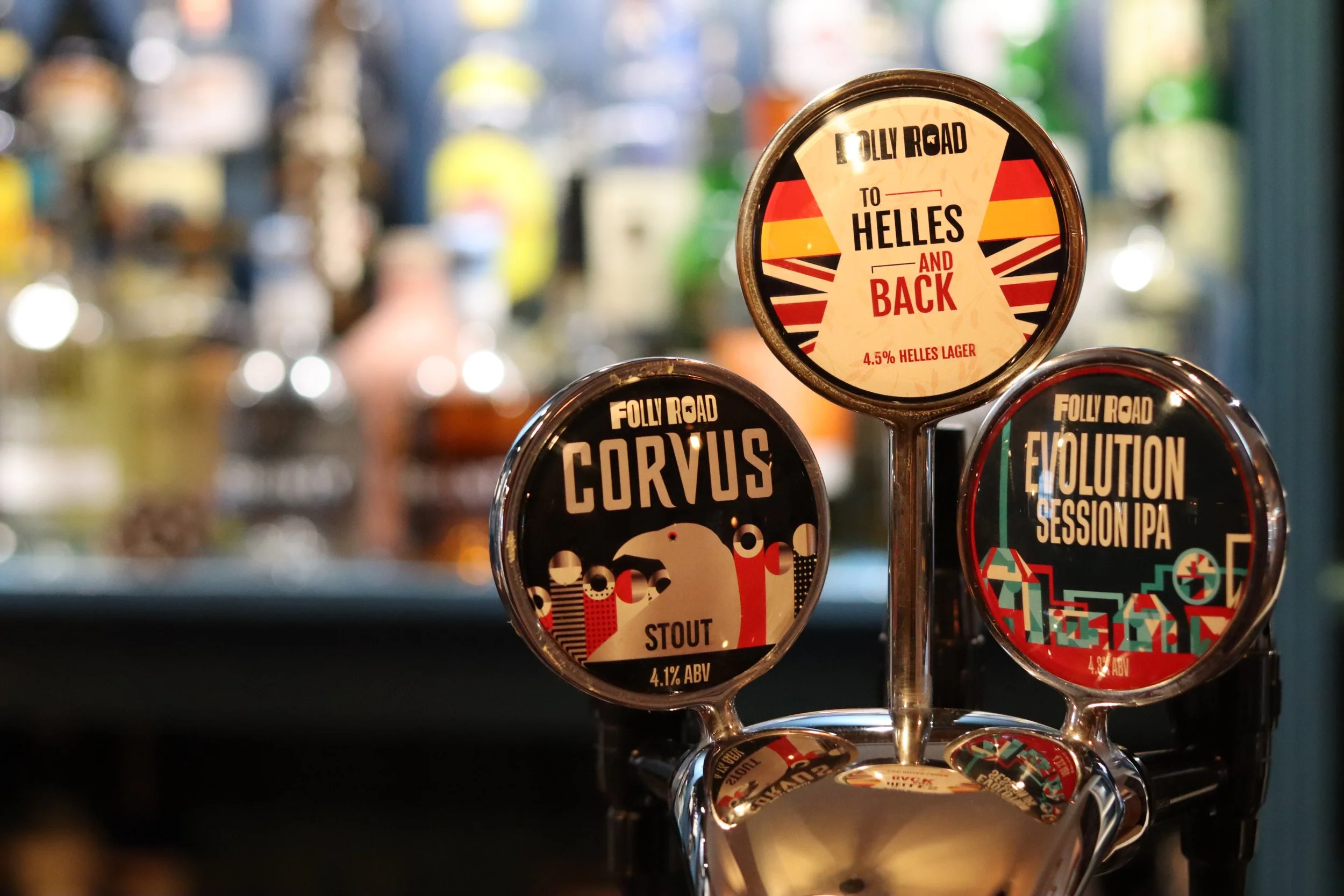 A tap handle display with three beer labels, featuring Folly Road beers: Helles and Back, Corvus Stout, and Evolution Session IPA, with a blurred background of a bar or brewery setting.
