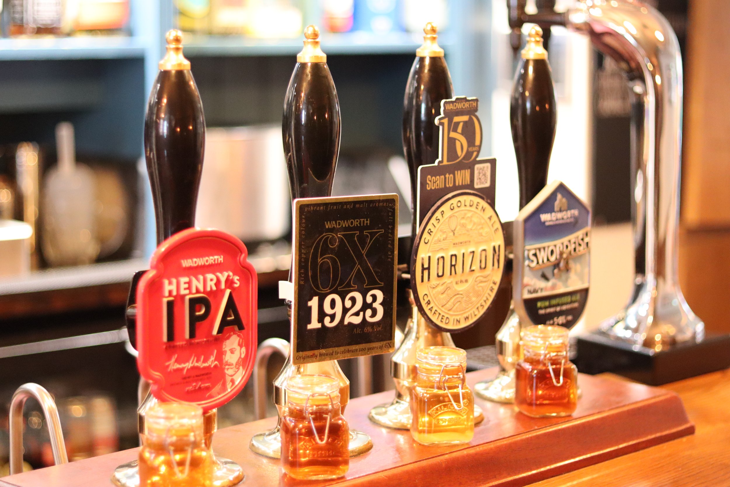 Four beer taps with unique labels at a bar, including Henley's IPA, 6X 1923, Horizon, and another with a festival coaster, with small jars of honey in front.