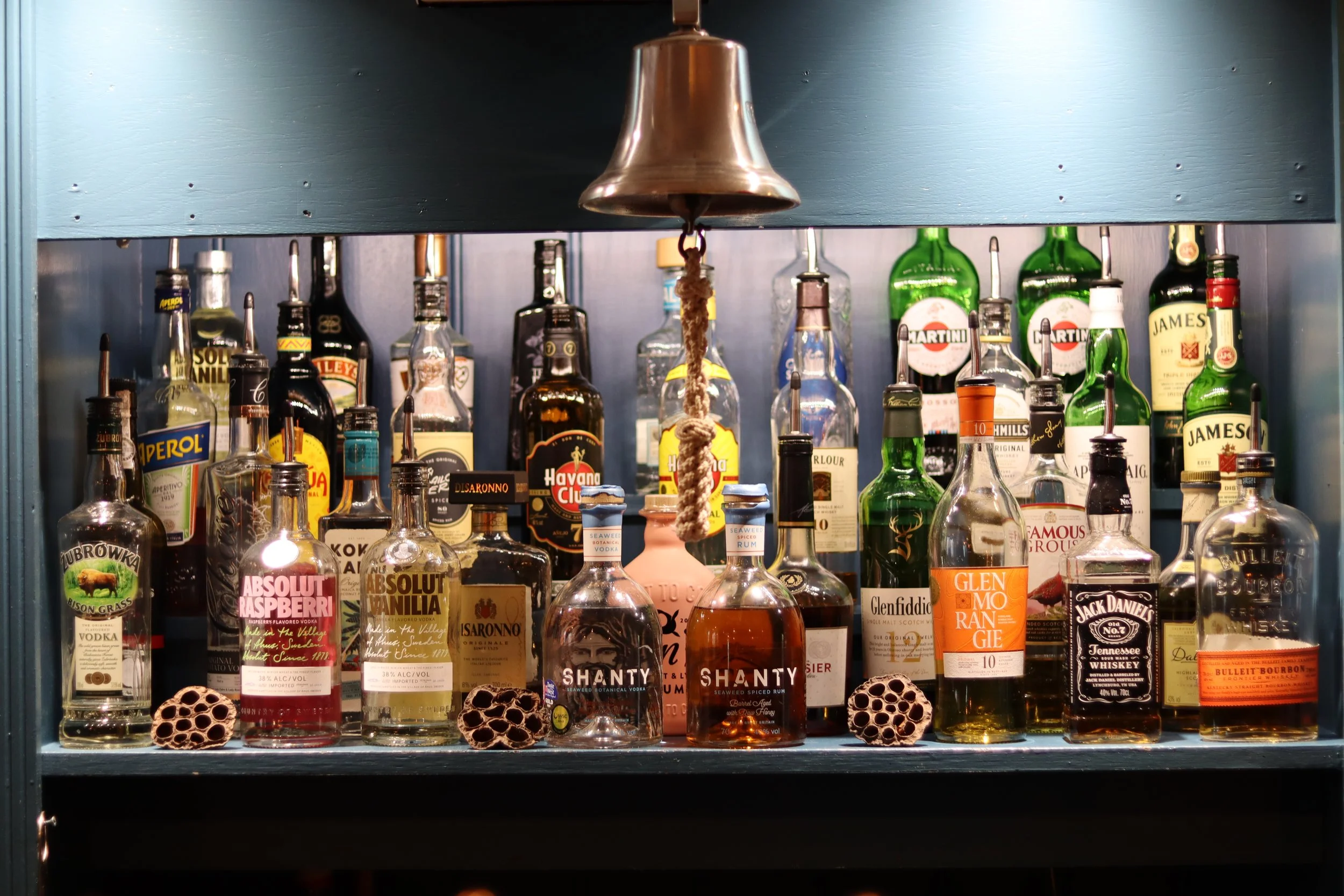 A collection of various liquor bottles displayed on a shelf in a bar, with hanging bell and decorative pods on the surface.