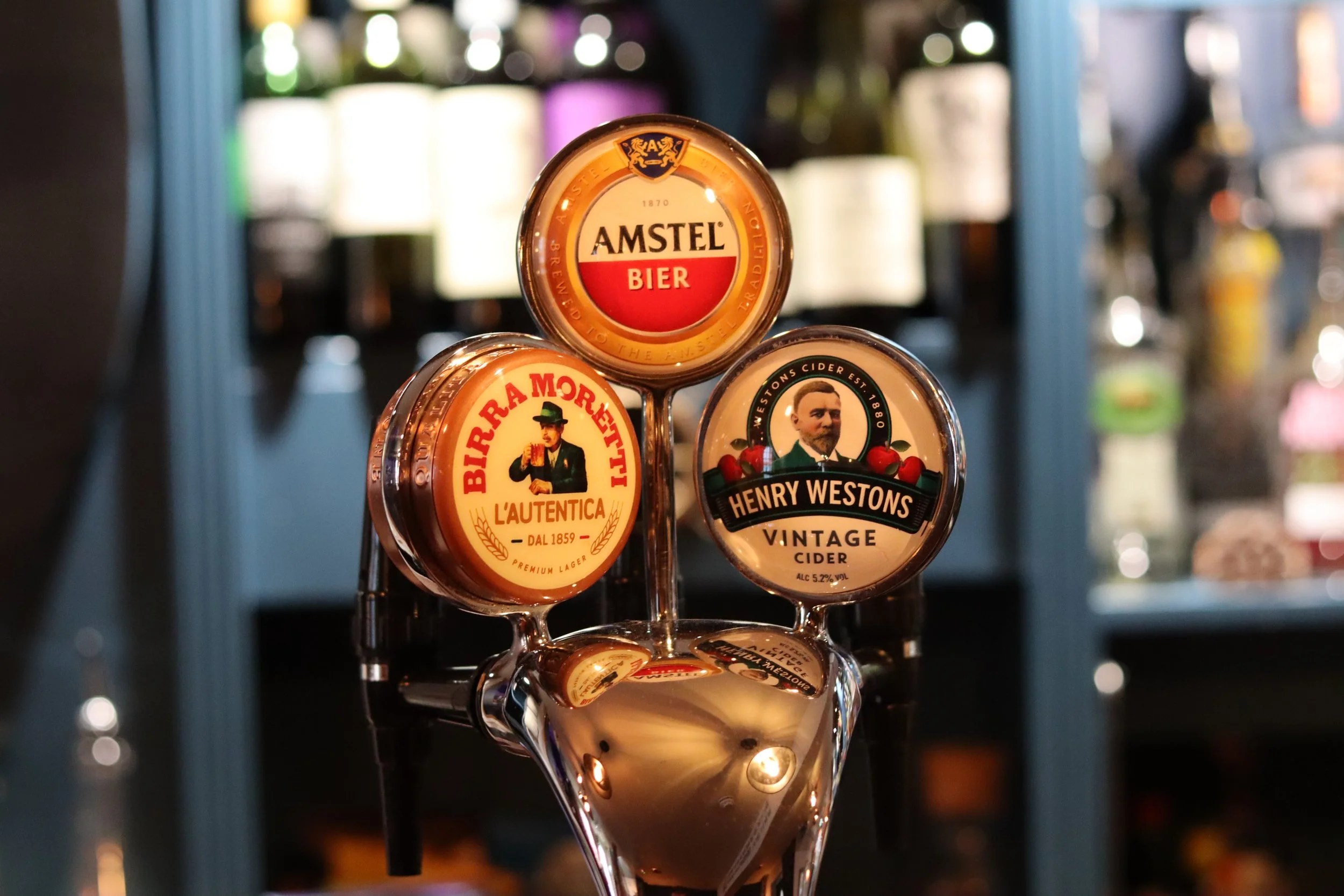 Three beer taps with different labels, featuring Amstel Bier, Birra Moretti L'Autentica, and Henry Westons Vintage Cider, in a bar setting with blurred bottles in the background.