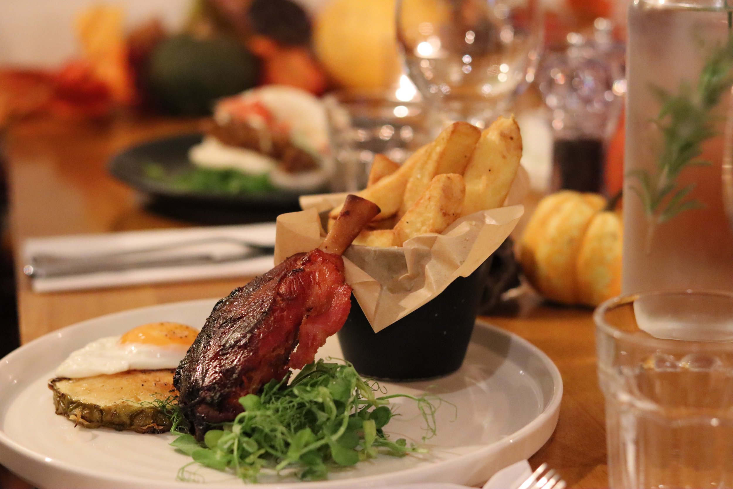 A plate with a baked potato topped with sour cream, a strip of crispy bacon, a fried egg, and some greens; a black cup of French fries in a small paper container; and a backdrop of a dining table with glasses, vegetables, and pumpkins.