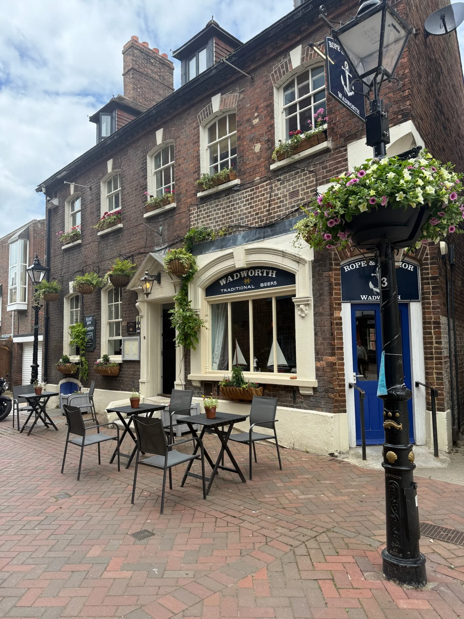 Outdoor seating area of a pub called WADWORTH with tables, chairs, and potted plants, located on a brick sidewalk beside a brick building with hanging flower baskets and a blue door.