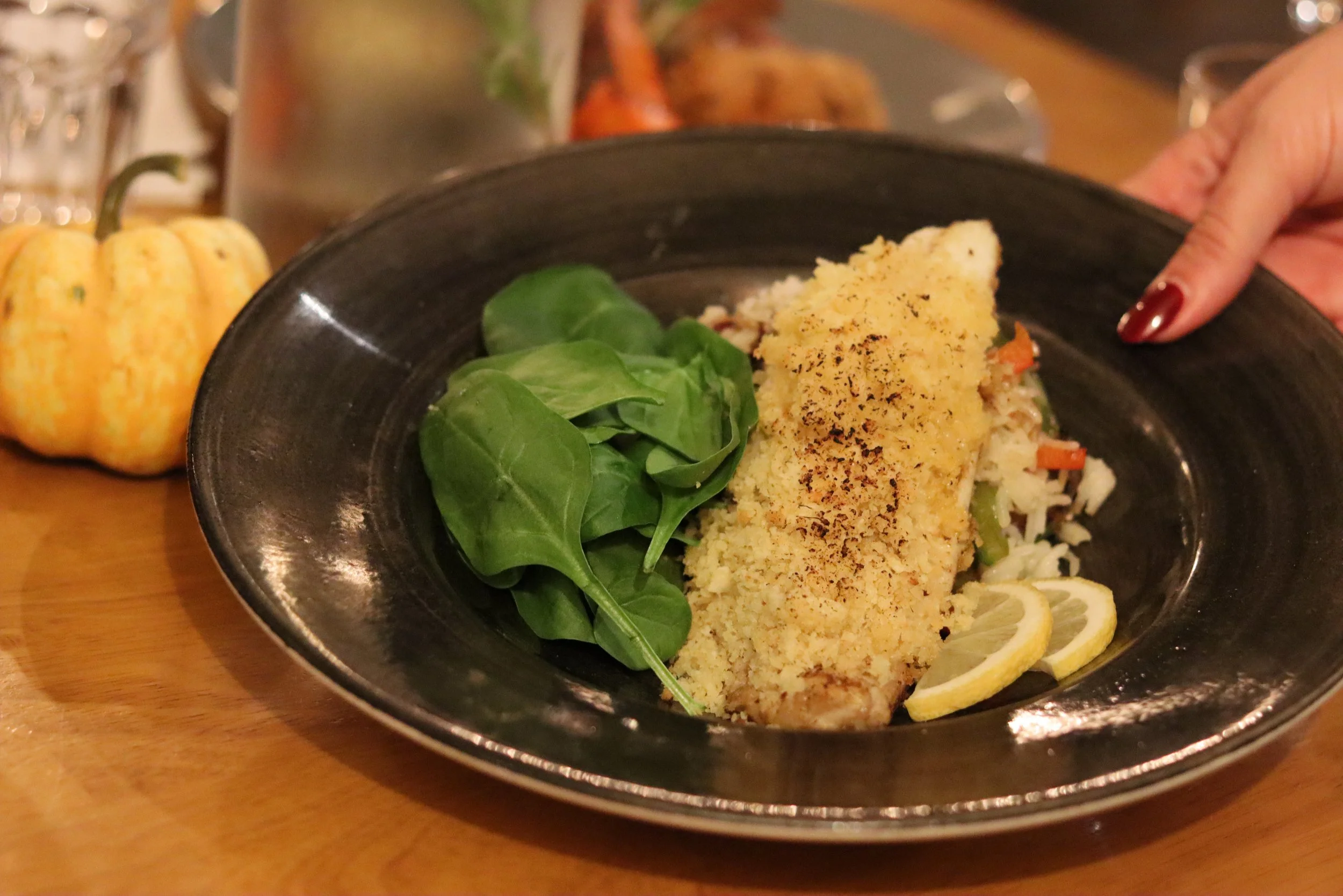 A plate of breaded baked fish fillet with lemon slices, green leafy vegetables, and vegetables in a black bowl. A small pumpkin decoration and a silver pot are in the background.