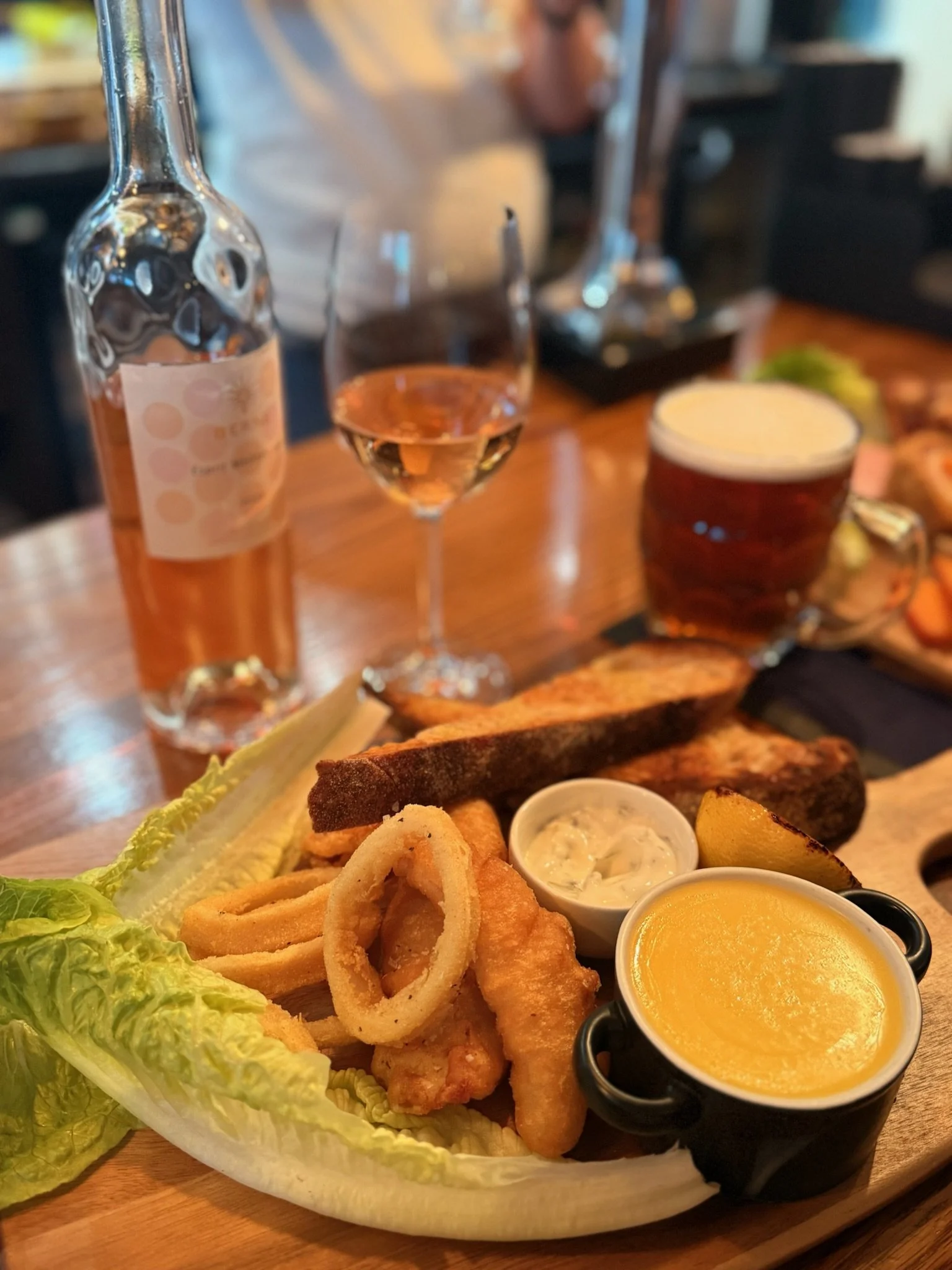 A wooden platter with fried seafood, onion rings, French fries, and dipping sauces, accompanied by a wedge of lettuce, served with a glass of pink wine, a bottle of rosé, and a glass of beer on a wooden table in a restaurant setting.
