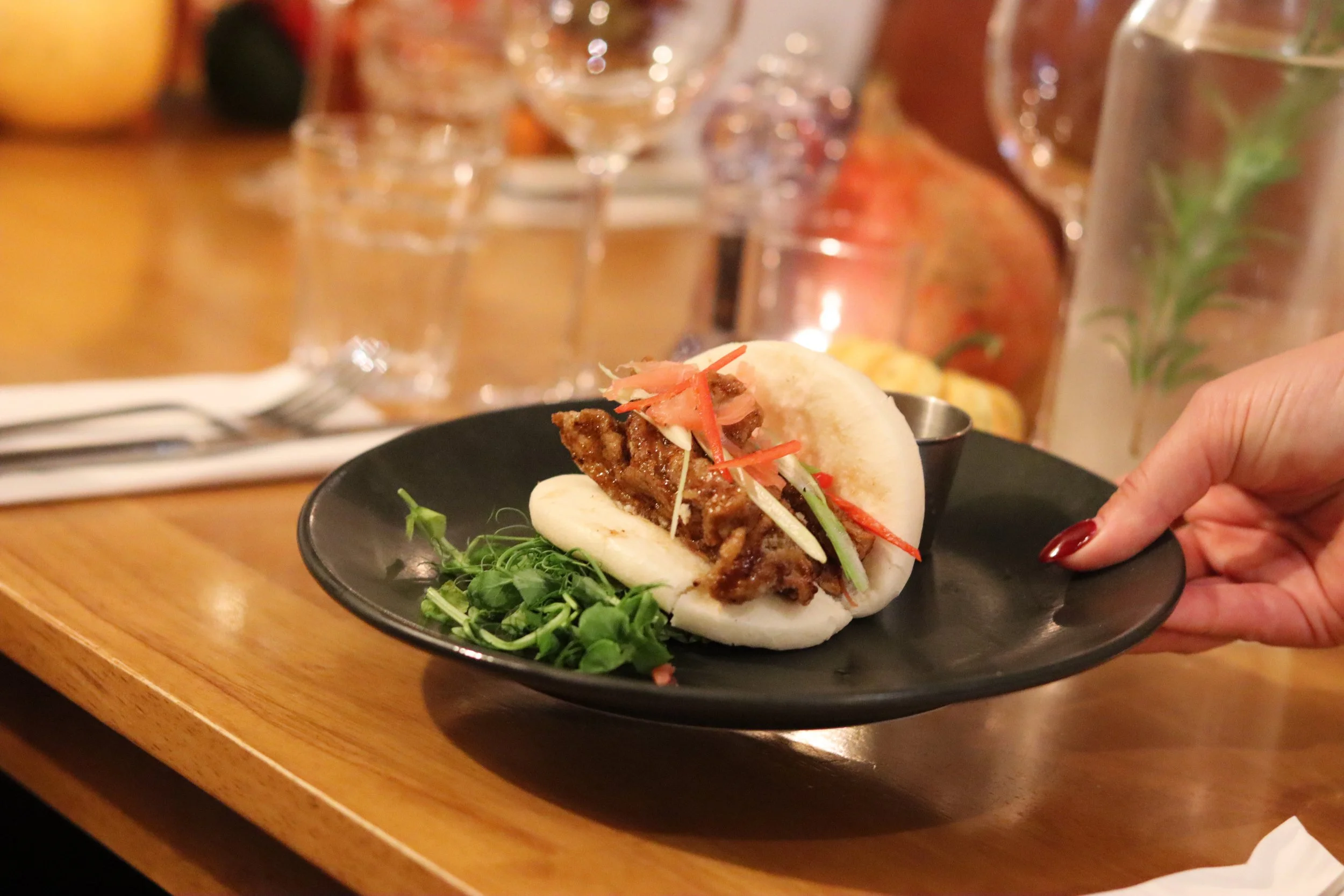A steamed bao bun filled with grilled meat, garnished with sliced vegetables, served on a black plate with greens, in a restaurant setting.