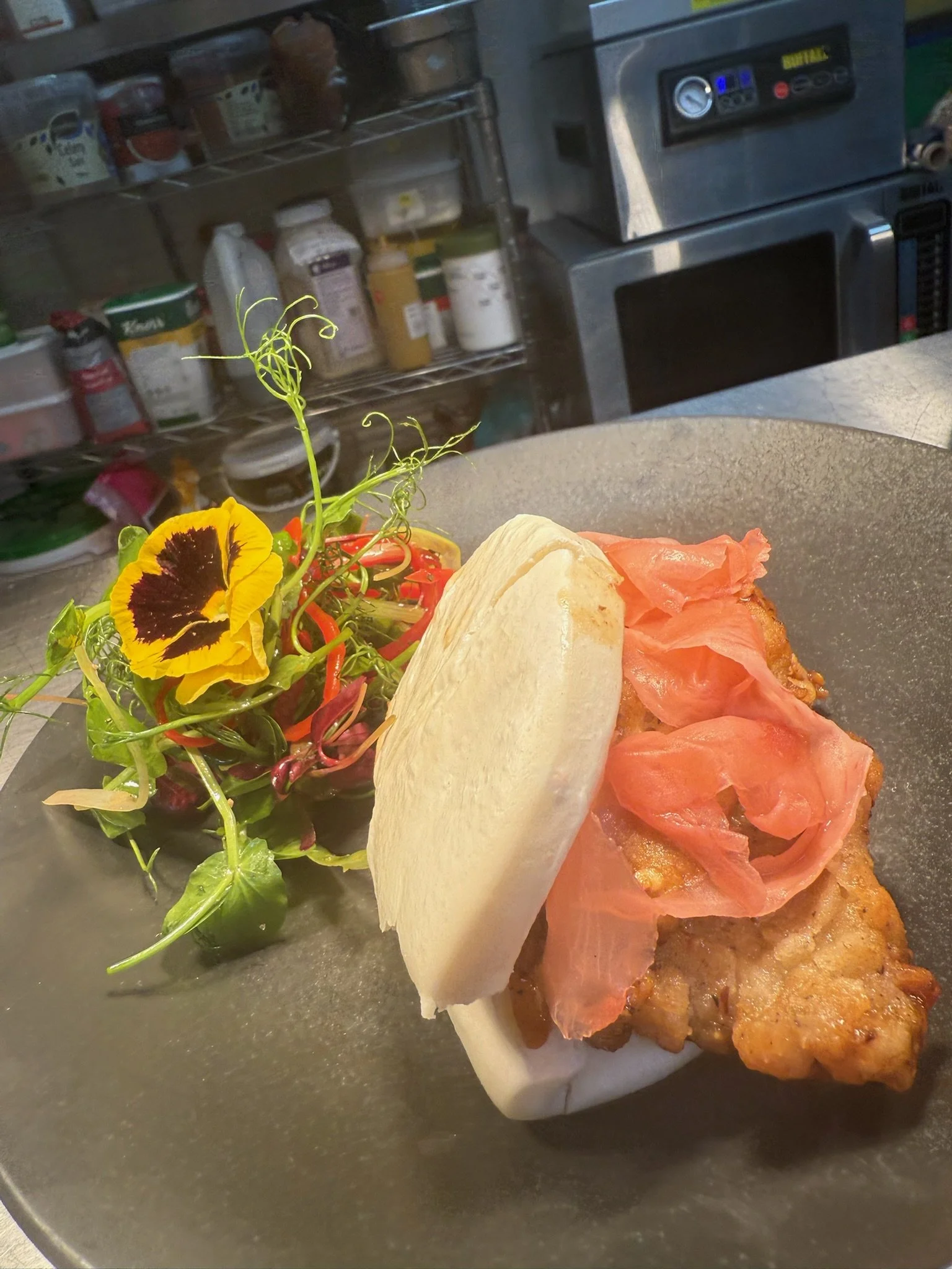 Fried chicken sandwich with pickled ginger, served with a side of mixed greens and edible flowers on a round black plate.