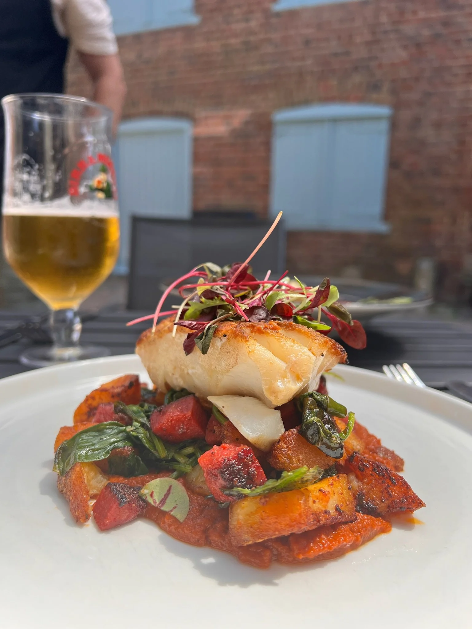 A plated dish with fish on top of roasted vegetables and tomato sauce, garnished with microgreens, with a glass of beer in the background, set outdoors against a brick wall.