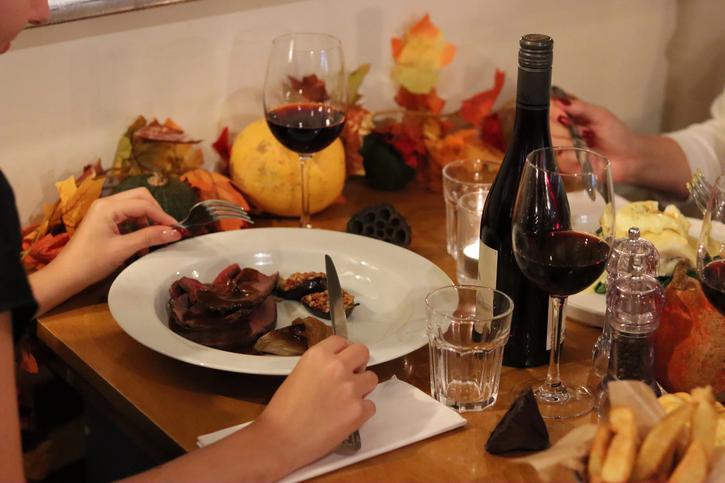 A family dinner table with wine glasses, a bottle of red wine, and plates of food, including roasted vegetables and sliced meat, decorated with autumn leaves and pumpkins.