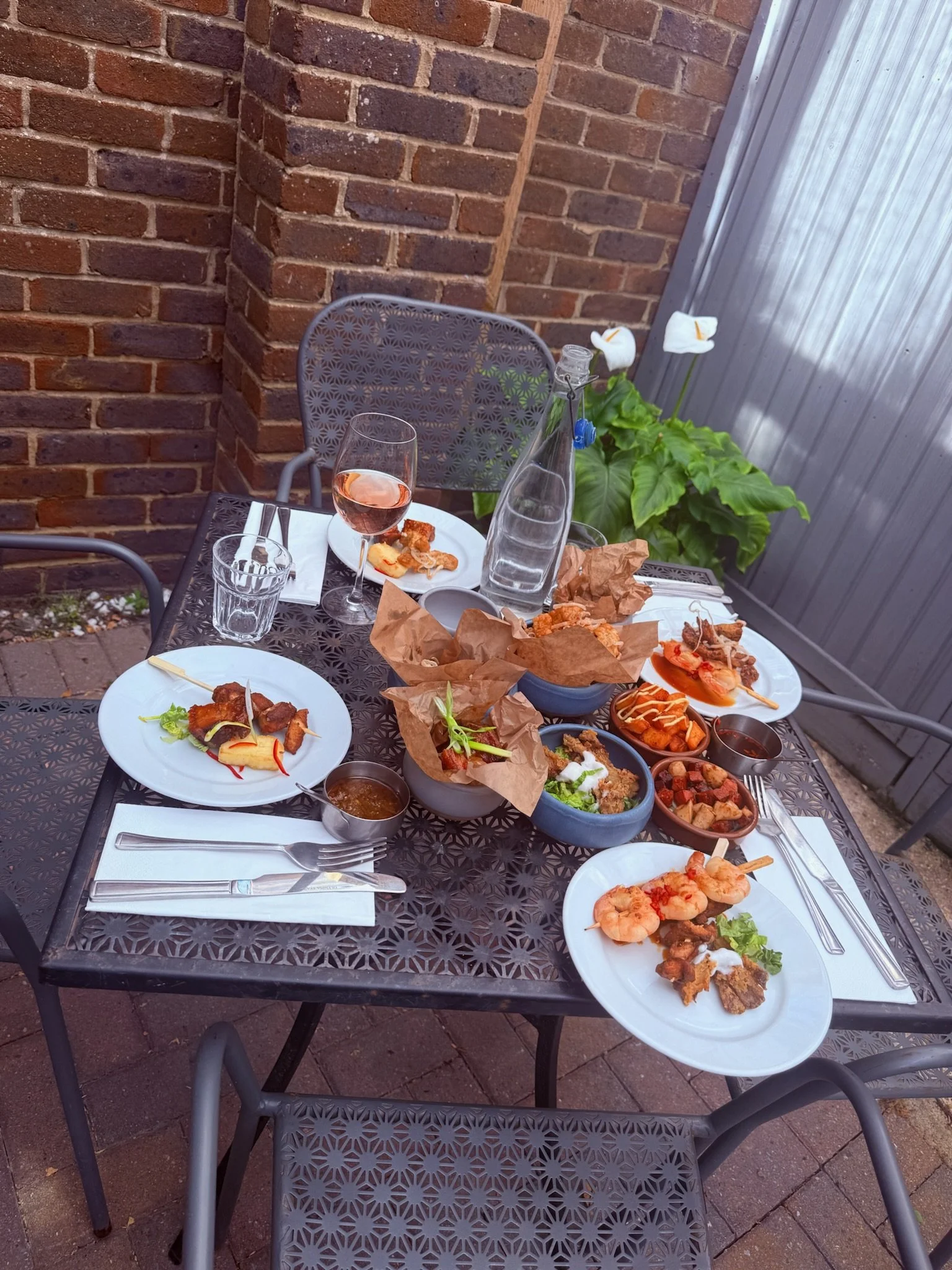 Outdoor dining table with various dishes and drinks, including a glass of rosé wine, a water bottle, bowls of food, and plates with skewers, set against a brick wall and metal fence.