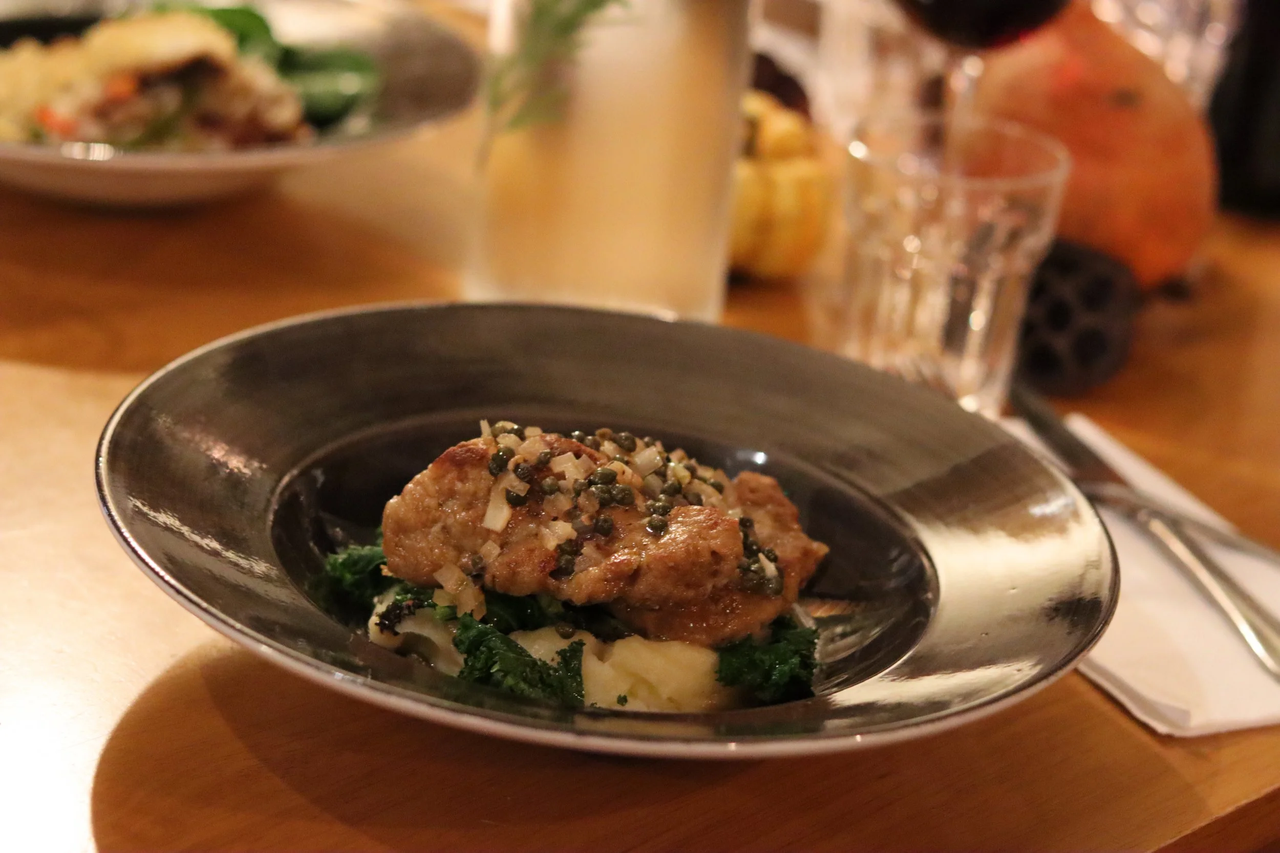 A plated entrée featuring a cooked meat patty topped with diced onion and herbs, served on a bed of greens and mashed potatoes, on a dark oval plate.