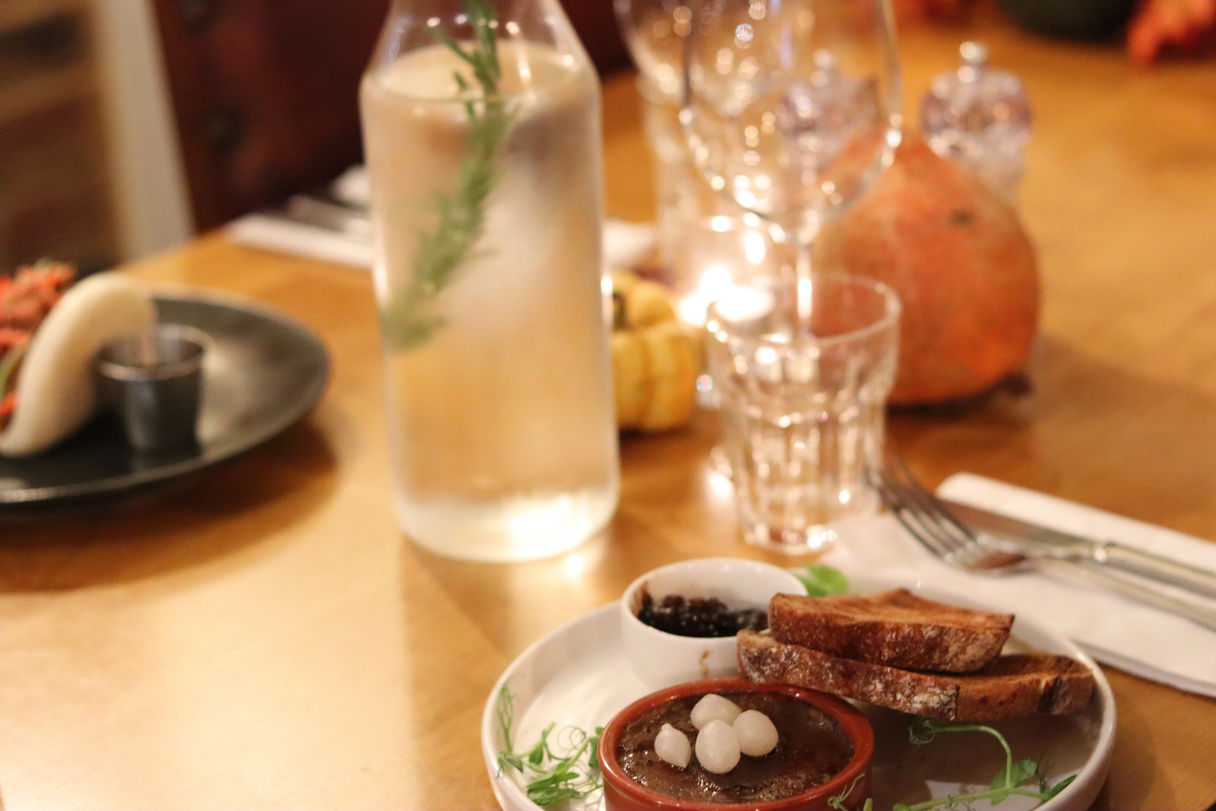 A table setting with a glass of water garnished with a sprig of herbs, a small plate with toasted bread slices, small bowls of jam and butter, and a background with a pomegranate, wine glasses, and a plate with tacos, all on a wooden table.