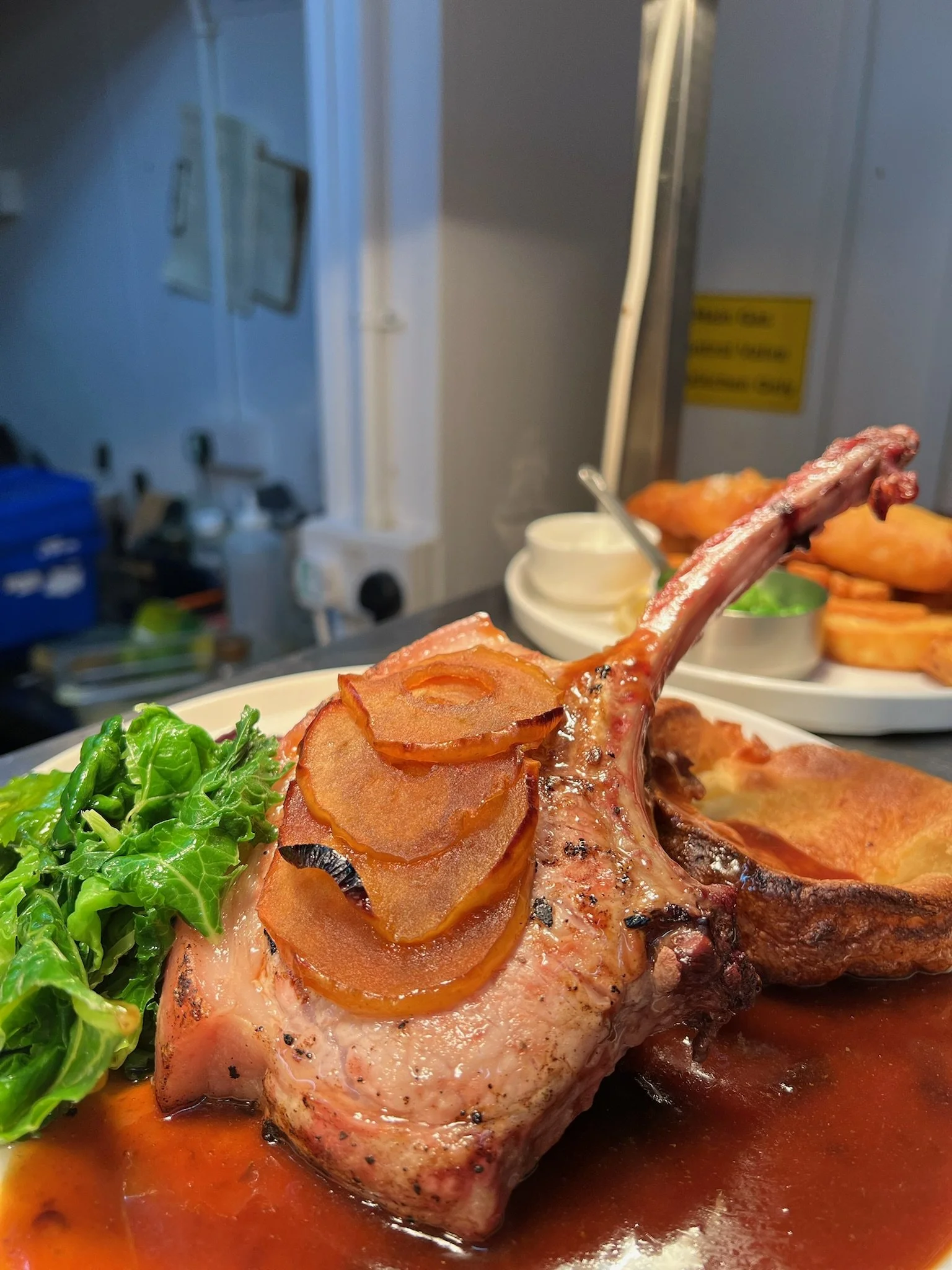 Close-up of a cooked lamb chop garnished with fried onions, served with a side of green leafy vegetables and a brown sauce on a white plate.