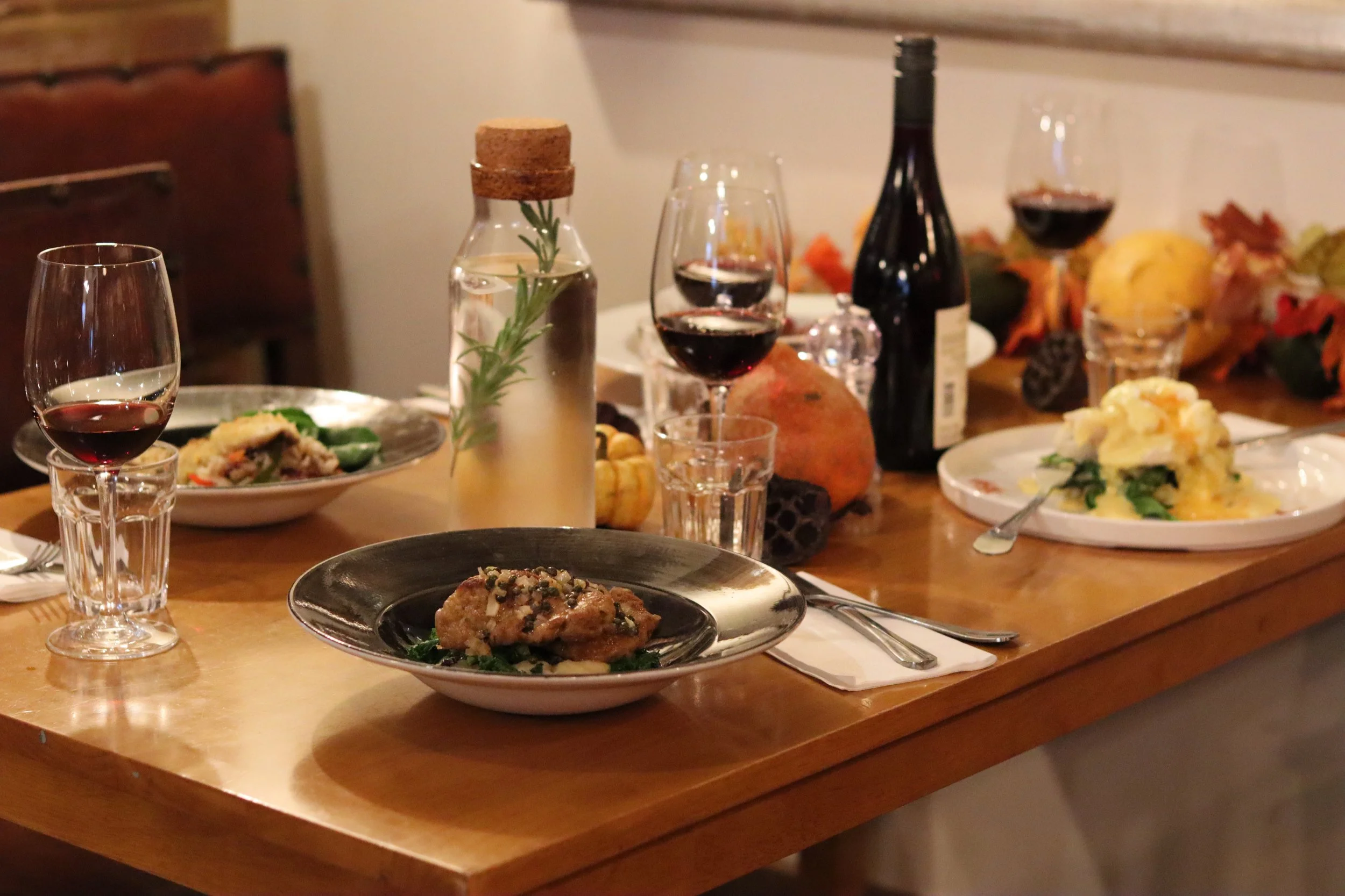 A wooden dining table set with plates of food, glasses of red wine, a bottle of wine, a decanter with herbs, and decorative autumn leaves and pumpkins.
